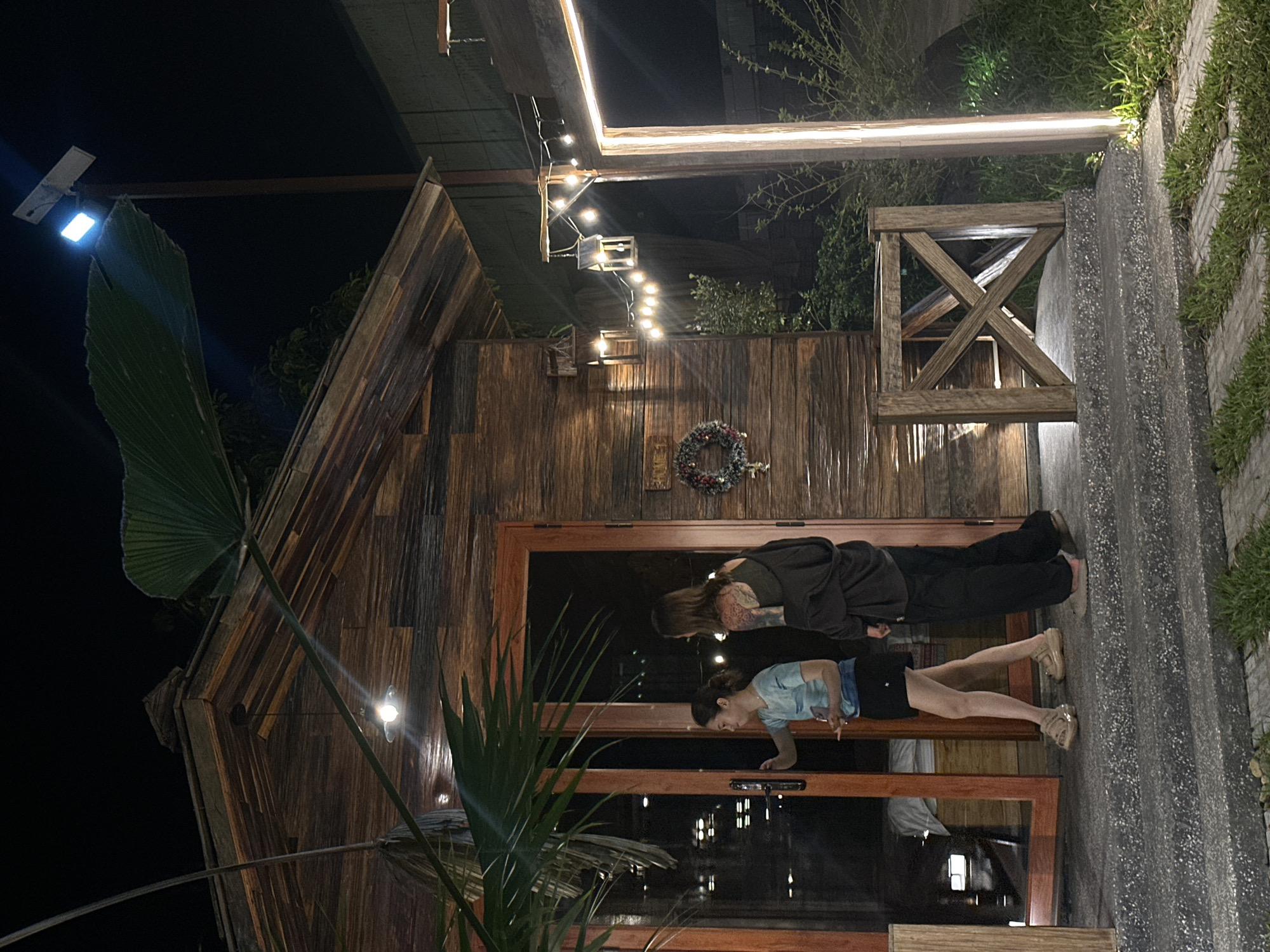The image depicts a nighttime outdoor scene, claimed to be in Nghia Lo Ward, Vietnam, featuring a rustic wooden building and two individuals. The building, constructed from horizontally laid wooden planks of varying shades, has a peaked roof and a prominent wooden-framed glass door, which is partially ajar. A small, round, downward-facing light fixture hangs from the roof overhang above the door. To the right of the door, a circular wreath adorned with red berries and green foliage is affixed to the wooden wall. Further right, a string of warm-toned light bulbs hangs between a horizontal wooden beam and an unseen anchor, with two open-frame lantern-style light fixtures also suspended.
Two female individuals are positioned near the building's entrance. The individual on the left, facing right, wears a light-colored, possibly blue or teal, short-sleeved top with a patterned design, black shorts, and light-colored platform sandals. Her left hand is near the door handle, and her right hand holds a small, light object. The individual on the right, positioned slightly behind and to the right of the first, wears a dark, possibly black, tank top or camisole underneath an open, dark, long-sleeved jacket or cardigan, and dark trousers or pants with flat, dark shoes. This individual has extensive tattoos covering her left arm and upper back/shoulder. Both individuals are approaching or entering the building.
The foreground consists of concrete steps leading up to the building, with a short wooden railing featuring an 'X' pattern on the right side of the steps. To the right of the railing, a vertical strip light illuminates a paved pathway interspersed with grass. In the upper right background, a tall pole supports a bright blue-white street light with an attached solar panel. Various green palm leaves and other foliage are present, some partially obscuring the building and the sky. The overall scene is dark, illuminated primarily by the artificial light sources.