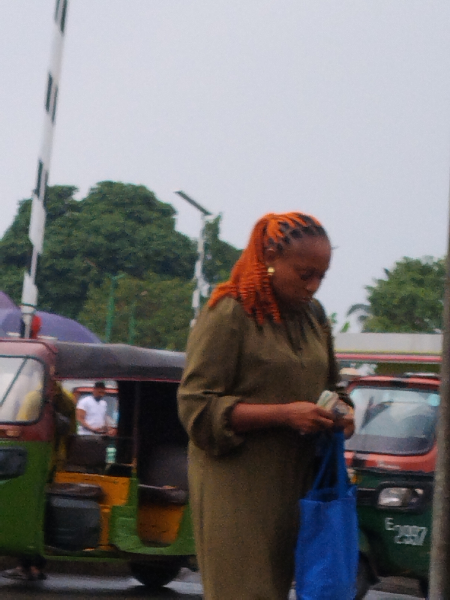 The image depicts a woman with orange braided hair standing on a street in Aba, Nigeria. She wears a long, olive-green dress and gold earrings, and holds a blue shopping bag and what appears to be money in her hands. Behind her, there are two motorized tricycle taxis, one in shades of green, yellow, and black, and the other in red and green. A person in a white shirt is visible inside the green tricycle. A white and black gate, possibly a barrier, stands to the left of the woman. Trees and overcast skies are visible in the background, along with street lighting. The license plate of the red tricycle reads "E-2037".