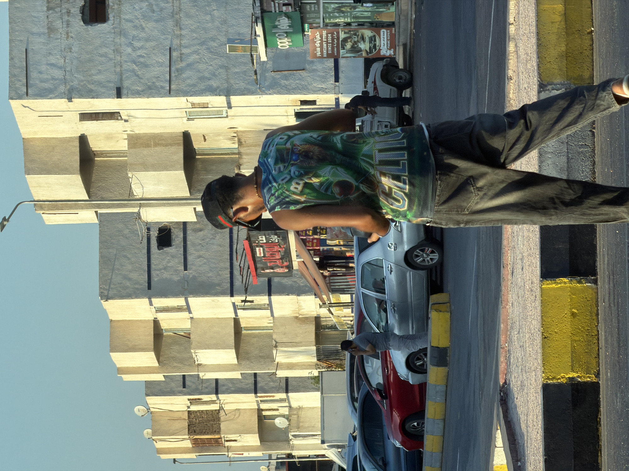 In Sharm El Sheikh, Egypt, a young man wearing a dark baseball cap and a sleeveless t-shirt with a green graphic design of a leprechaun in a basketball uniform and the word "CELTICS" is walking away from the camera. He is wearing dark, baggy jeans and white shoes. He holds a dark object, possibly a phone, in his left hand, bringing it up to his ear as if having a conversation. The man is walking on a paved street in front of a row of light-colored buildings with a modern, somewhat stark architectural style featuring geometric projections and balconies. Several cars are parked or driving in the background, including a red sedan, a silver sedan, and a white pickup truck. A man in a gray t-shirt and pants is visible near the silver sedan, also appearing to be on his phone. Signs with Arabic text and advertisements for businesses like "OPPO" and "HAMZAWY STORE SINCE 2004" are present on the buildings. The sky is a clear, pale blue, indicating daylight, and the scene is illuminated by sunlight casting shadows, suggesting it is late afternoon or early morning. Yellow and black concrete barriers line the edge of the street.