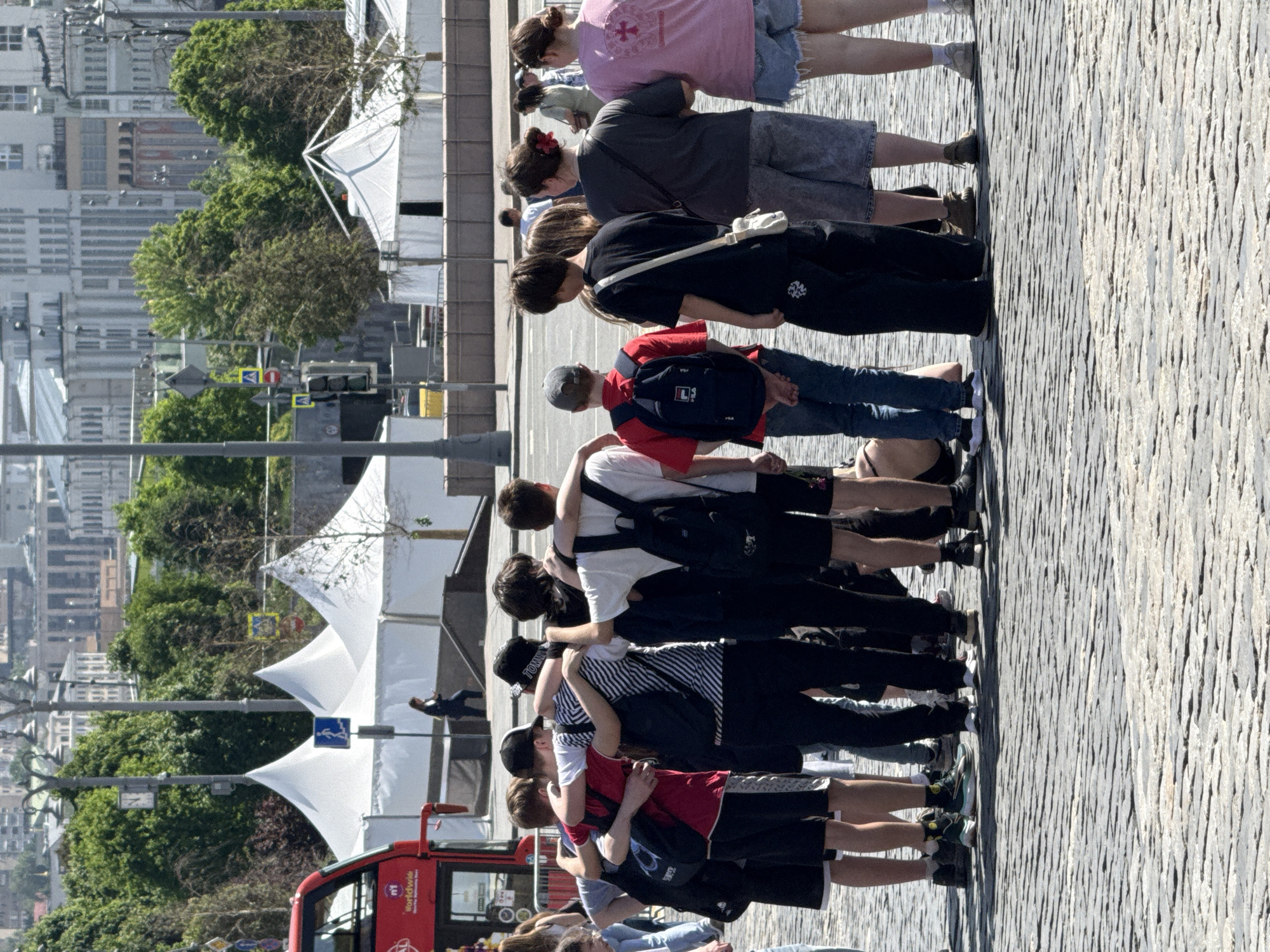 A group of approximately a dozen people, mostly young adults, stands close together in a line on a cobblestone-paved public area. The individuals are facing the same direction, with arms around each other, suggesting a group activity or organized event.
Behind the group, there are white tents set up, indicating a possible festival or public gathering. Beyond the tents, there are trees and buildings, including what appears to be an observation tower, suggesting an urban environment. The presence of a red double-decker bus partially visible in the lower left corner of the image suggests the location is near tourist attractions in an unidentified city. A man is shown walking on an elevated platform toward the city. Streetlights with pedestrian crossing signs are seen between the tents.