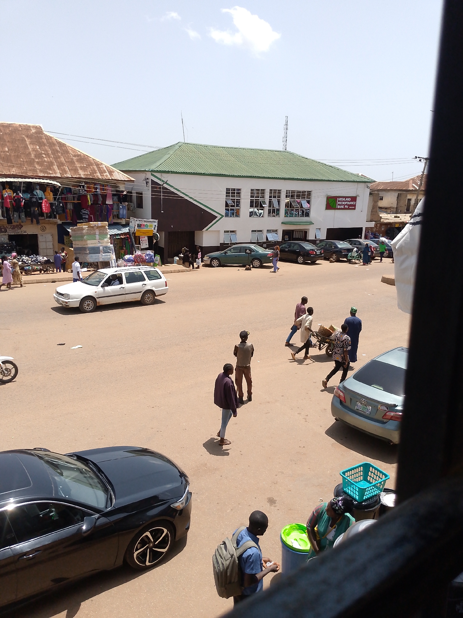 The image depicts a bustling street scene in Bukuru, Nigeria, likely during the daytime under a clear, sunny sky. The foreground is dominated by a black sedan parked on the right side of the road. In the mid-ground, several people are walking and interacting. A white station wagon is driving on the street, and a few other cars are parked or moving in the background.
On the left side of the street, market stalls are set up, displaying clothing and other goods. Several people are browsing or walking past these stalls. A Highland Microfinance Bank PIc building with a green roof is visible in the background, along with other buildings characteristic of the area.
A group of men are pushing a cart loaded with baskets, suggesting commercial activity or the transport of goods. The overall atmosphere is one of everyday life and commerce, with people engaged in various activities on the street and in the market. The lighting suggests it is midday.