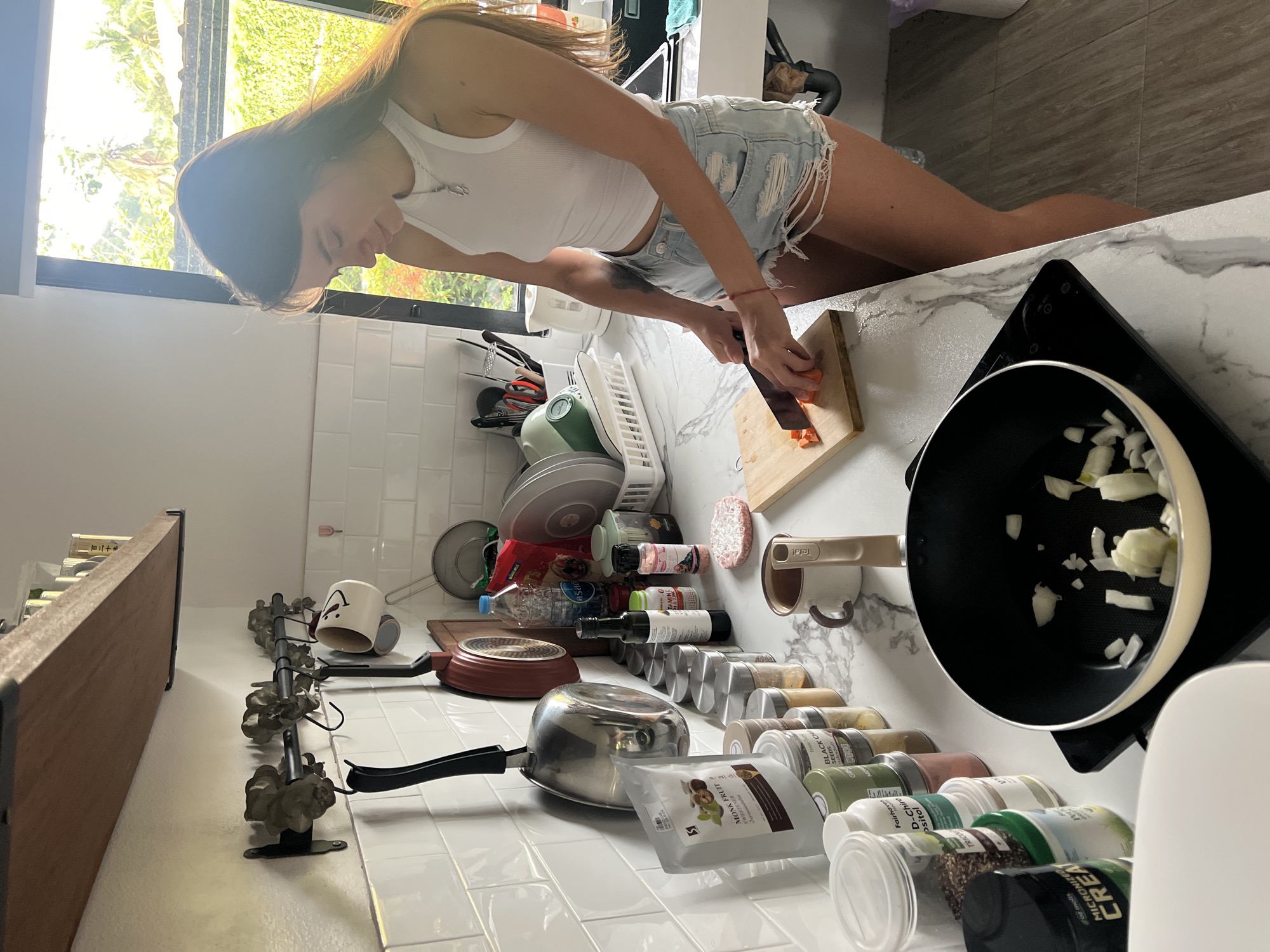 A woman wearing a white tank top and ripped denim shorts is cutting a carrot on a wooden cutting board on a white countertop in a kitchen. A black frying pan containing chopped onions sits on a black induction cooktop to the right of the cutting board. To the left are various kitchen items including a stainless steel saucepan, spice jars, a red frying pan, plates, and a dish rack. A window is visible in the background. The location is บ้านบางรักษ์, Thailand.