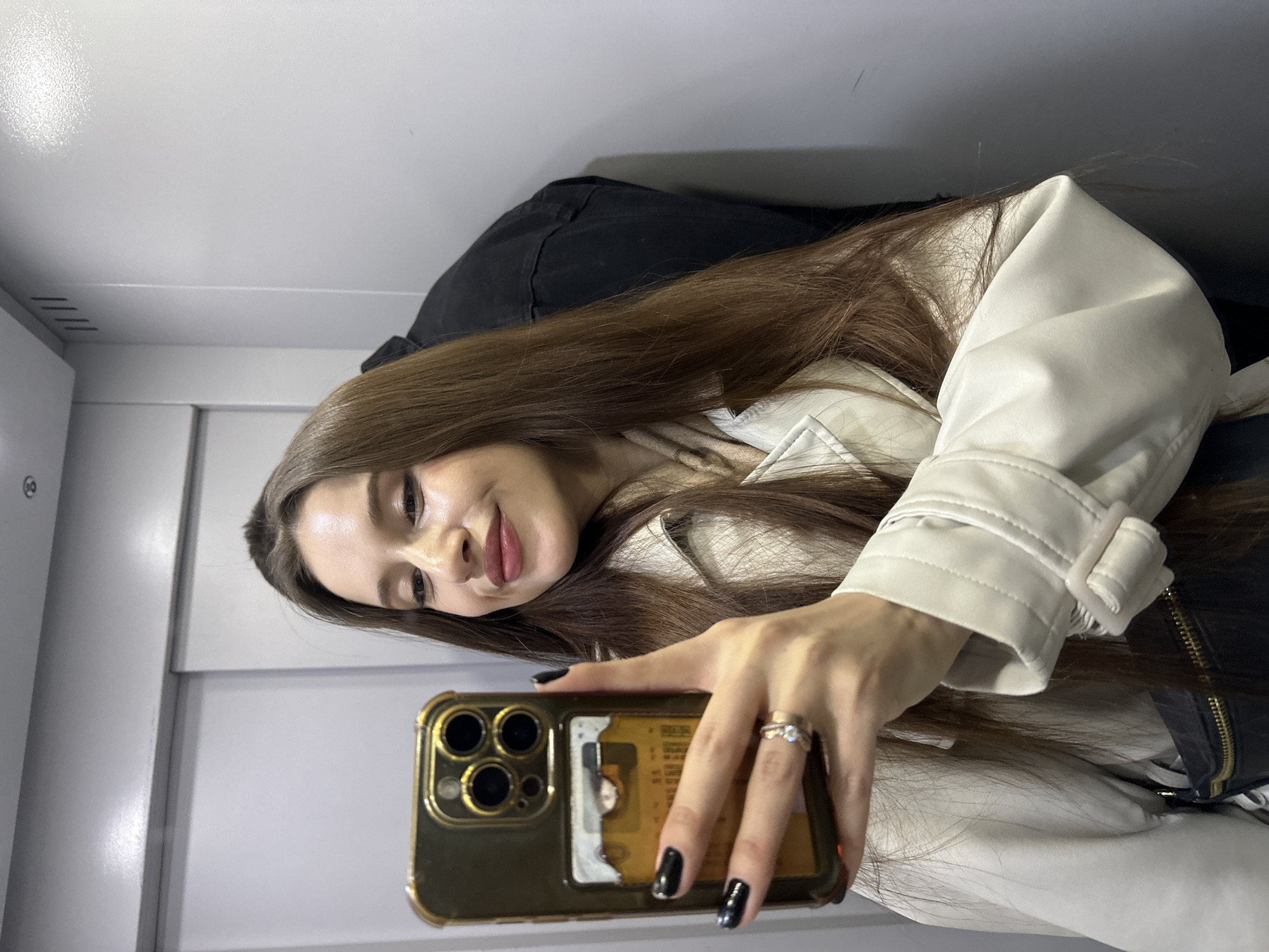 A young woman is captured in a mirror selfie, holding an iPhone-style smartphone with a gold case in her left hand. She has long, straight, dark brown hair and a light complexion, looking towards the camera with a soft smile. Her hand, prominent in the foreground, features black nail polish and a ring on her left ring finger.
She is wearing a light beige or off-white collared jacket with a visible white buckle on its cuff. Behind her, a dark garment or object is partially seen. The background consists of plain, light-colored walls, suggesting an indoor setting such as an elevator or a minimalist room. There are subtle vertical seams in the wall panels and some horizontal grooves on the left side of the frame. A card with red and white markings is tucked into the back of the phone's transparent gold case. The lighting is bright and even. No legible text is visible in the image.