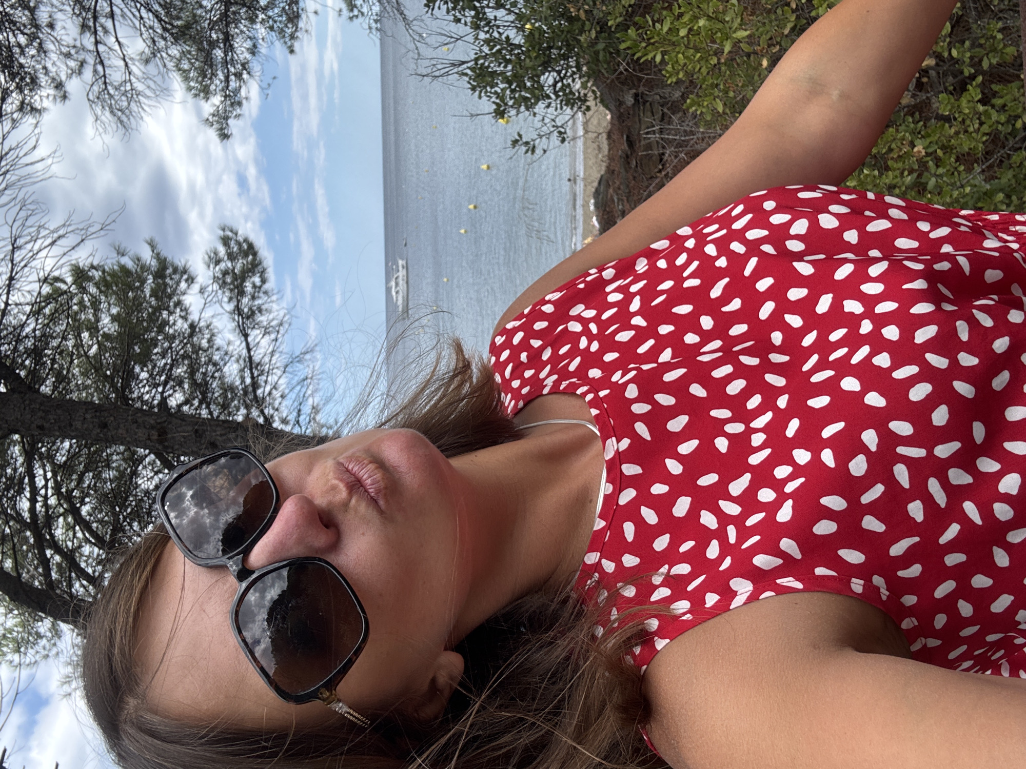 A woman is taking a selfie on a sunny day by the sea. She is wearing large, dark sunglasses and a red dress with white polka dots. Her lips are puckered as if she is blowing a kiss. In the background, the sea is calm with a few buoys and a white boat. There are trees and bushes on either side of the frame, suggesting an outdoor setting, possibly a park or a coastal path in Port-Vendres, France. The sky is partly cloudy with blue patches.