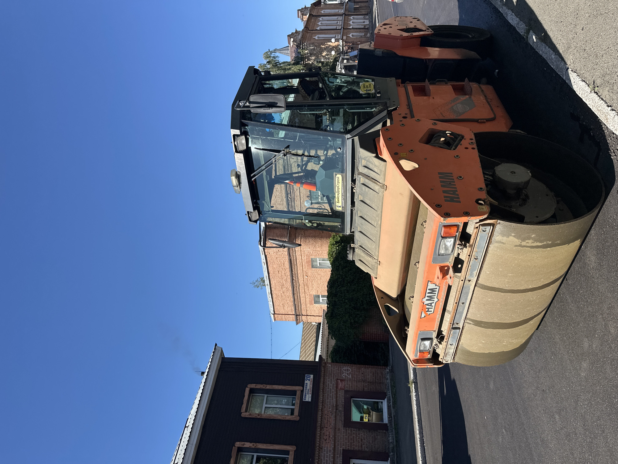 In Biysk, Russia, on a clear day with a bright blue sky, an orange HAMM vibratory roller is positioned on newly laid asphalt. The roller, a piece of construction equipment, is the primary subject of the image. Its large, dirt-covered drum is in the foreground, and its cabin with tinted windows is visible behind it. Two yellow stickers are affixed to the cabin: one with Cyrillic text that appears to be "БийскДорСтрой" and another with the same text and a smaller graphic.
To the left of the roller, a two-story building with a dark wooden facade and brick lower section is visible. A street sign on this building reads "Советская 20" in Cyrillic, indicating the location is Sovetskaya Street, number 20. A thin plume of smoke rises from the chimney of this building. Adjacent to it, a brick building with multiple windows is visible, partially obscured by dense green foliage. Further in the background, another brick building with ornate architectural details can be seen. The scene appears to be a street undergoing resurfacing or construction, with fresh asphalt in the foreground and a shadow cast by the roller on the road. There are no people clearly visible in the immediate vicinity of the roller, though there might be a figure inside the cabin, obscured by the reflections and tint of the glass.