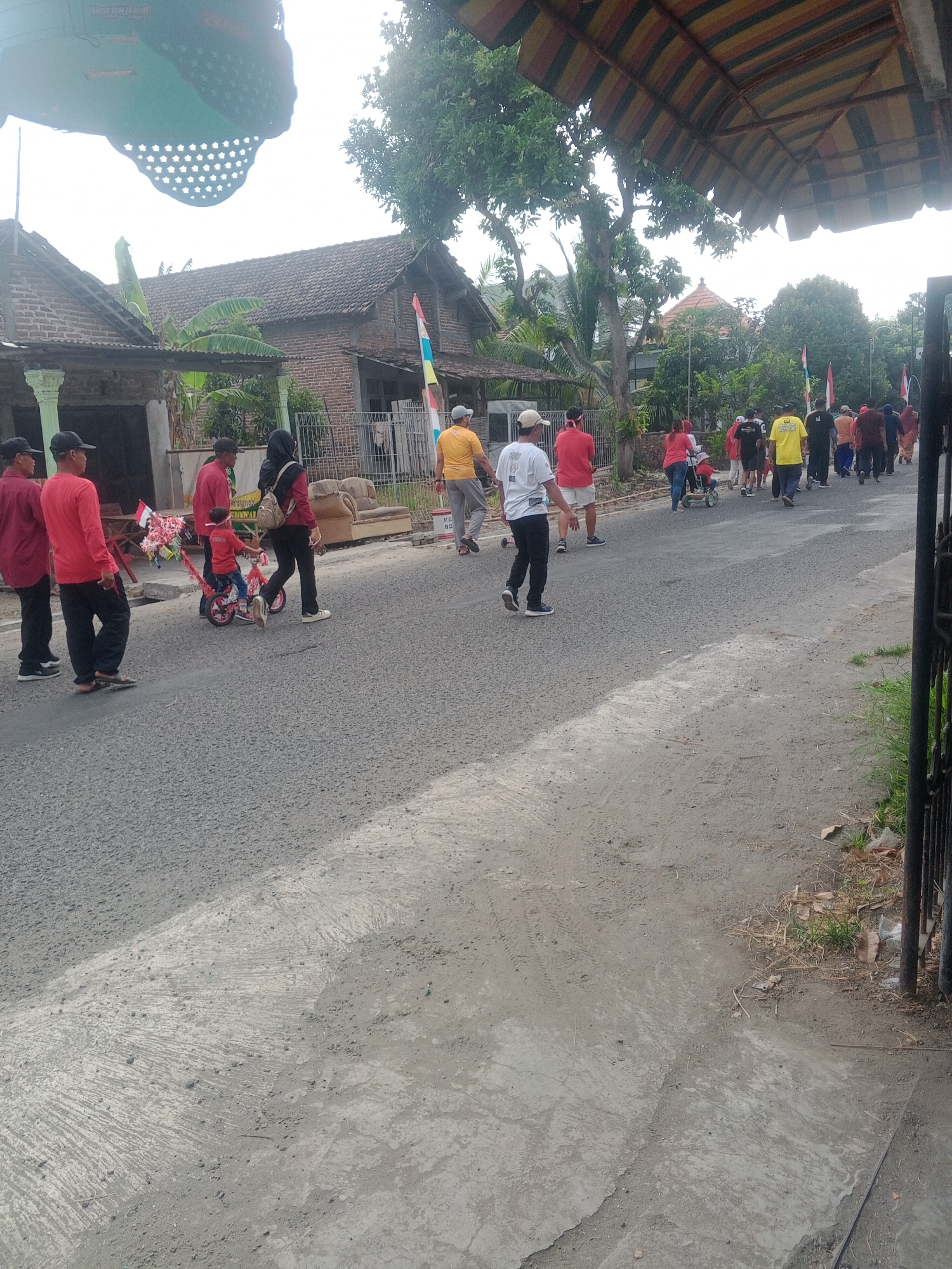 The image shows a street scene in Lembah, Indonesia, likely during a community event or procession. Several people are walking on the asphalt road, some in casual attire, others in matching shirts, suggesting participation in an organized activity. A child is riding a small tricycle adorned with decorations, possibly a float or part of a parade. On the left side of the road, houses with traditional Indonesian architecture are visible, characterized by tiled roofs and brick walls. Trees and foliage line the street, indicating a somewhat rural or suburban setting. The sky is overcast, suggesting a cloudy day. A dark metal fence runs along the right edge of the image, bordering the road and a patch of dry grass and leaves. The overall impression is one of a lively local gathering taking place in a residential area.