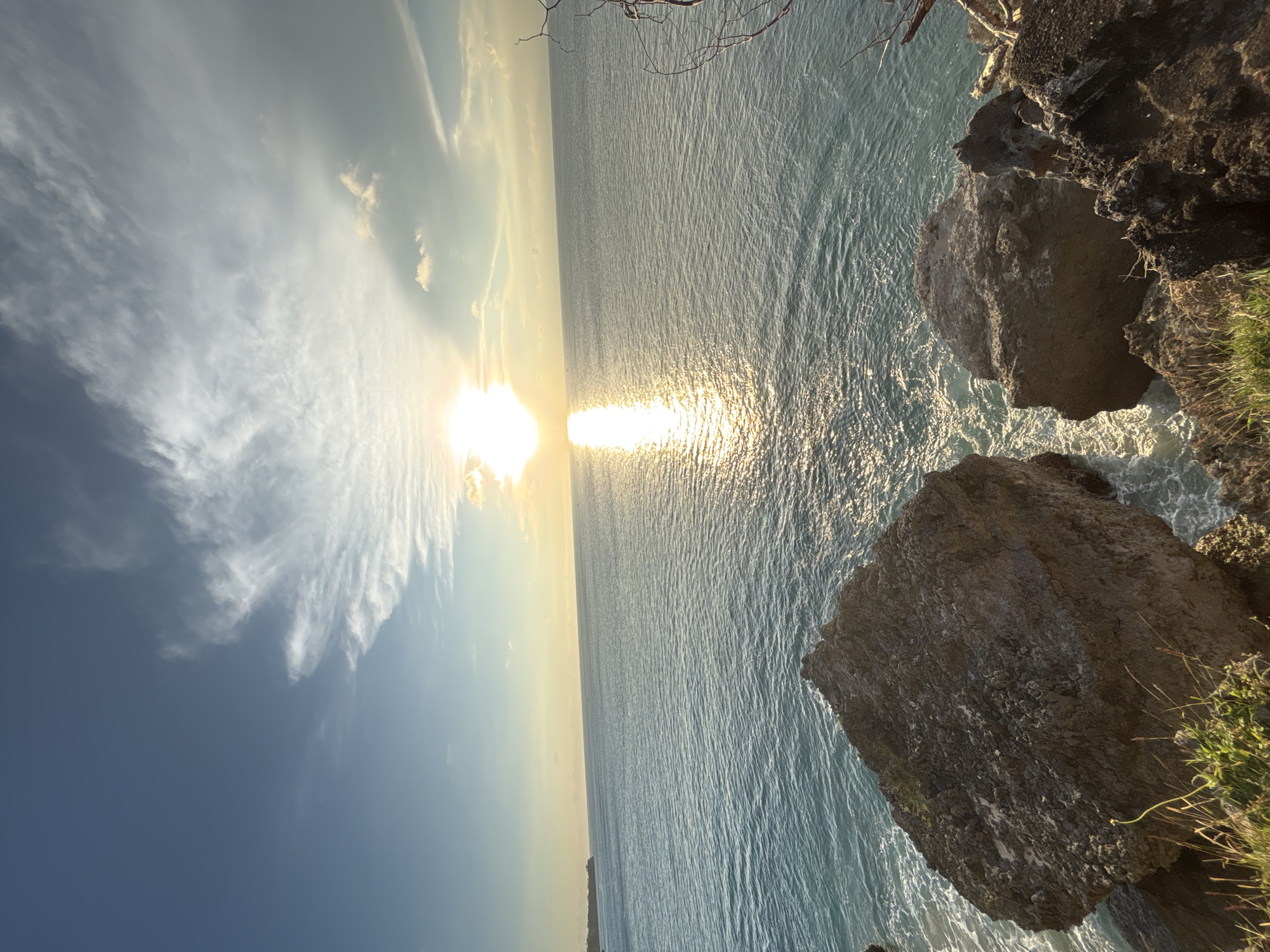 The image shows a seaside view from a cliff. The sun is setting over the water, creating a shimmering reflection. Rocks are visible in the foreground on the right side, with some vegetation growing around them. The sky has clouds, and the lighting suggests either sunrise or sunset. Based on the request parameters, it is located at Kuta Selatan, Indonesia, but no specific landmark is readily apparent in the provided image.