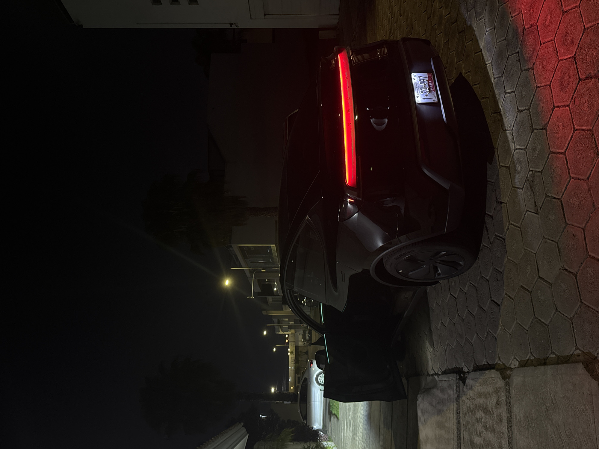 The image shows a dark-colored SUV parked on a hexagonal-tiled driveway at night in San Bernardino Tlaxcalancingo, Mexico. The vehicle's rear passenger-side door is ajar. The SUV's red tail light is illuminated, and the license plate reads "SZ-997". A red glow is cast onto the driveway tiles near the vehicle. In the background, streetlights illuminate a residential street with other parked cars and buildings. A white wall is visible in the foreground. The focus is primarily on the parked SUV and its immediate surroundings.