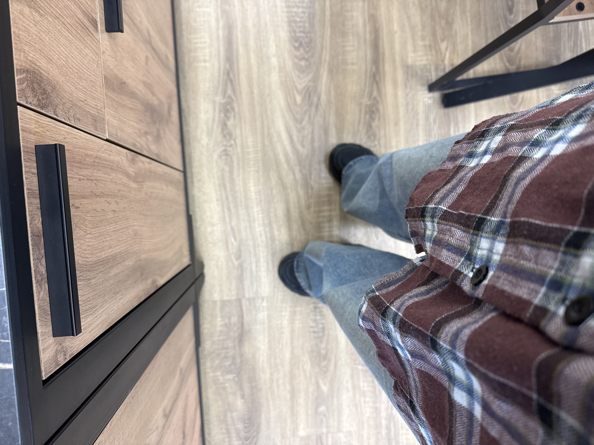The image shows a first-person perspective, looking down at a person standing on a light-colored wooden floor. The person is wearing blue jeans and dark shoes. A brown and maroon plaid flannel shirt is visible. To the left is a wooden cabinet with black handles. In the upper right corner, part of a black metal table leg is visible. The exact location within the city of Chaikovsky, Russia, cannot be determined from the given image.