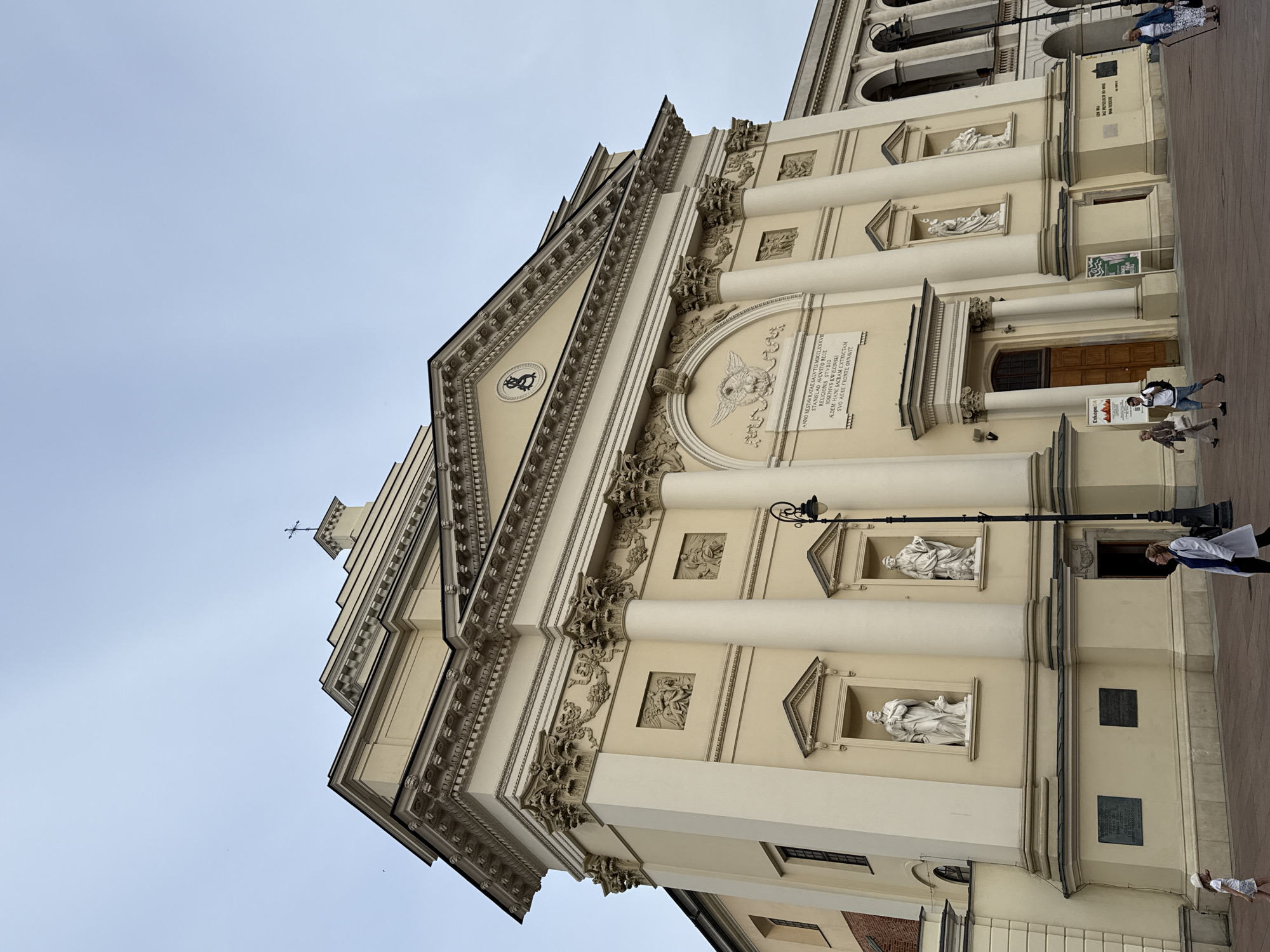 This is a low-angle, vertical shot of a pale yellow neoclassical building, likely a church, against a pale, overcast sky. The building features a prominent triangular pediment adorned with decorative carvings and a round medallion with a winged figure. A large inscription is visible on the facade. Flanking the entrance are large, fluted columns, and above them, niches contain white statues.
Several people are present on the pavement in front of the building. One woman in a white coat and blue scarf walks away from the camera towards the entrance. Another woman, wearing a white dress and hat, is looking at the building from a distance, to the left of the frame. Two individuals are closer to the bottom right, one with a backpack. The pavement is a reddish-brown color, indicating it's likely cobblestone or brick. A black lamppost stands near the upper levels of the building.
The inscription on the building appears to be in Latin. The text "ANNO RESTAVRATAE SALVTIS MDCCLXXXVE" suggests the year 1785. Another line reads "STANISLAD AVGVSTO REGE," indicating King Stanislaus Augustus. The words "RELIGIONIS STUDIO JOSEPHUS KWIECIN SKI" likely refer to a religious dedication and the name of the individual who commissioned or funded the construction.
The overall impression is one of historical architecture and a public space, possibly a city square, on a quiet, overcast day. The angle of the shot emphasizes the grandeur of the building.