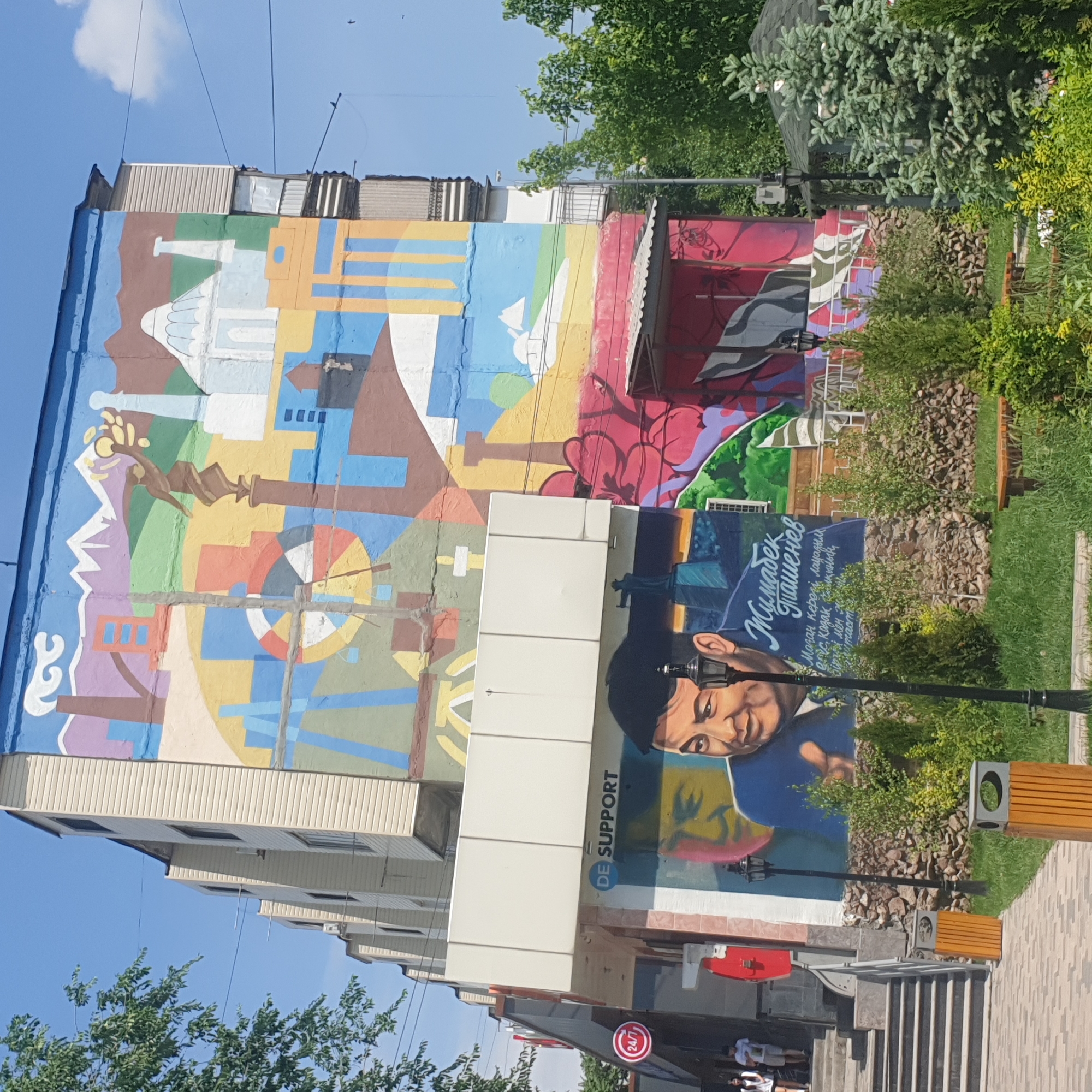 The image depicts a colorful building facade with two distinct sections. The upper section features a vibrant, abstract mural composed of various geometric shapes and architectural elements, rendered in a wide range of colors. Below this mural, a more realistic portrait of a man in a suit and cap is painted, accompanied by text in a Cyrillic script. The sign "DE SUPPORT" is visible between the two sections.
In the foreground, there's a street scene with benches, street lamps, landscaping, and a staircase leading up to the building. A "24/7" sign is visible on a business below the mural. People are partially visible at the bottom of the staircase. The mural is located in Shymkent, Kazakhstan.