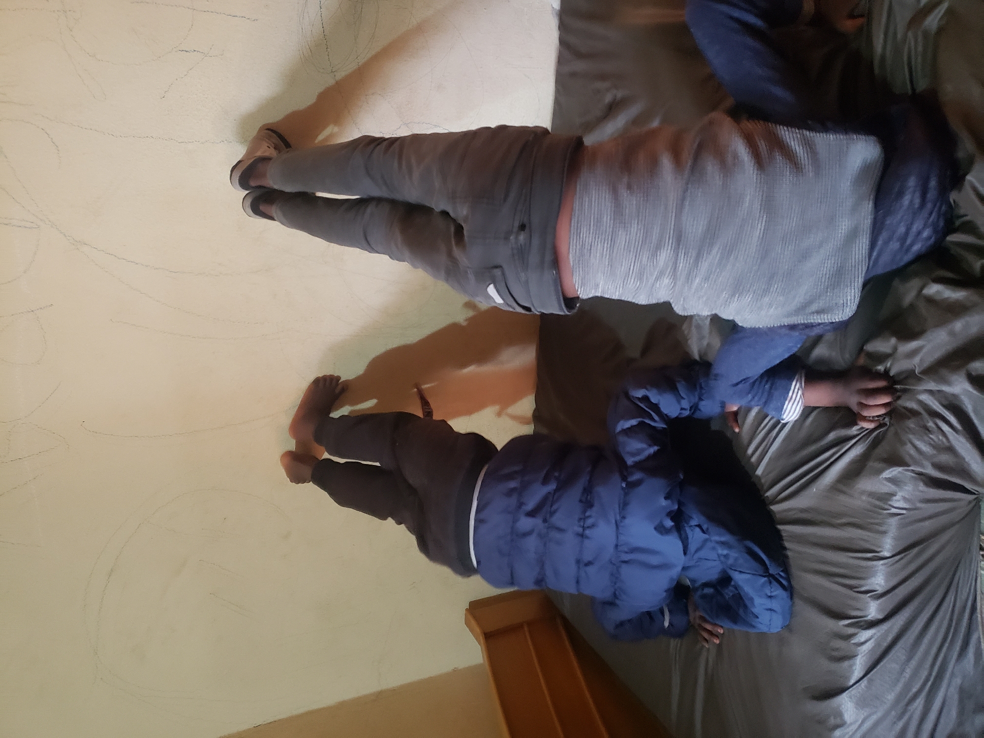 Two young children are performing handstands against a light-colored wall. The child on the left is wearing a dark blue puffer jacket and dark pants, with bare feet. The child on the right is wearing grey pants and a light grey striped shirt, with dark shoes. Both children are upside down with their legs extended upwards. They are positioned on a dark, possibly mattress-like surface. The wall behind them has faint pencil markings, suggesting it may be a drawing surface or has been drawn on. There is a wooden structure to the left of the frame, possibly a bed frame or shelf. The lighting appears to be indoor, casting shadows on the wall. The overall environment seems to be a room, possibly a bedroom or playroom.