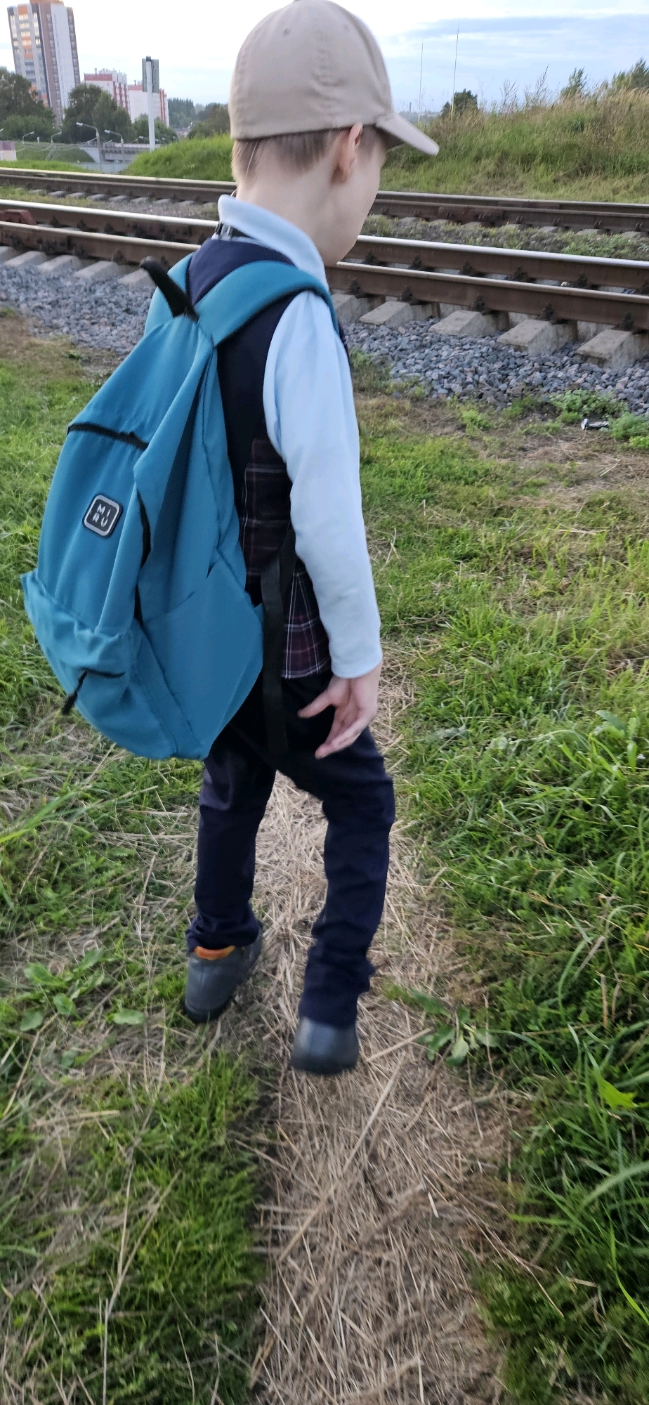 A child wearing a light blue long-sleeved shirt, a dark vest with a plaid pattern, dark pants, and a light brown baseball cap walks away from the camera on a dry, grassy path next to railroad tracks. The child has a teal backpack on their back. The railroad tracks run horizontally across the mid-ground of the image. In the background, there are trees and several tall buildings, some of which appear to be residential apartment complexes. The sky is overcast. The scene is likely a residential area adjacent to railway infrastructure.