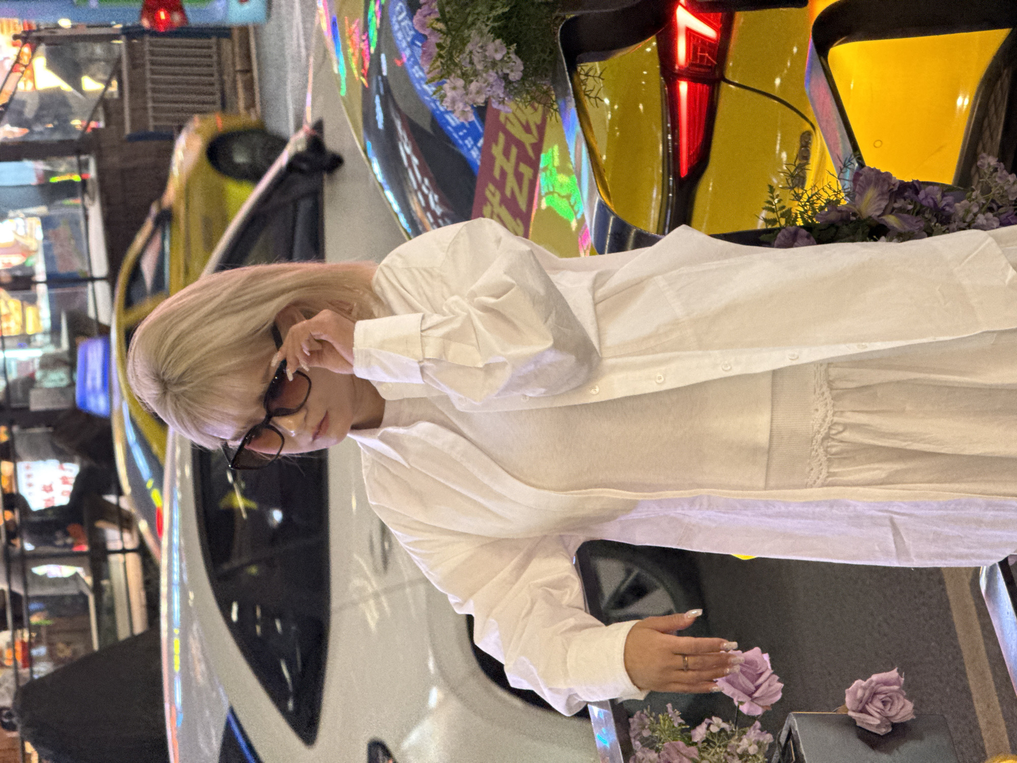 The image displays a woman standing on a city street at night in Liangjiang, China. The woman, positioned centrally, has blonde, shoulder-length hair and wears a white collared long-sleeve shirt over a white ribbed top and a white pleated skirt. She wears dark-framed sunglasses with brown-tinted lenses, her right hand is raised, touching the right temple of the sunglasses. A gold-colored ring is visible on her left ring finger, and her left forearm rests on a dark, reflective barrier.
The immediate foreground features two dark, rectangular planters containing purple and white floral arrangements, with two purple rose-like flowers hanging from the arrangement on the left. The background reveals a bustling urban street scene illuminated by various artificial lights. Multiple cars are visible: a white car partially obscured behind the woman, and two yellow cars, one directly to her right showing reflections of colorful neon signs and red taillights, and another further in the background to her left. Reflections of glowing signs, some with Chinese characters, are prominent on the car surfaces and across the cityscape. Blurred figures of pedestrians and brightly lit building facades line the street.