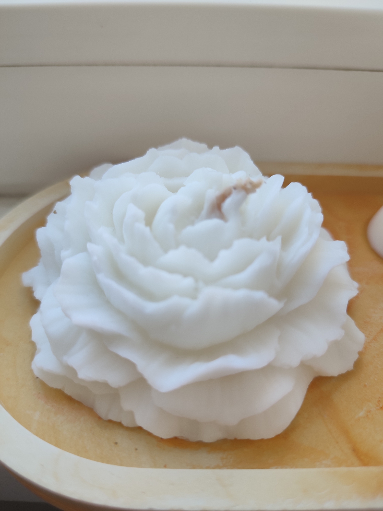 A white, rose-shaped candle sits on a light-colored wooden tray. The candle has many layered petals, and a small, brown wick is visible in the center. The tray is oval and has a slightly raised edge. The background is blurred and appears to be a light-colored surface, possibly a shelf or table. There are no people or other objects clearly visible. This image does not provide any information about its location in the city of Voronezh, Russia.