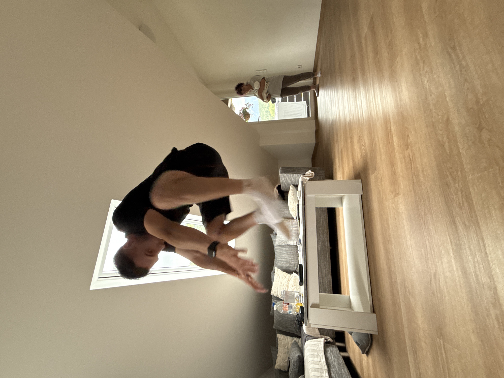 The image depicts an indoor scene, likely a living room, with a man in mid-air, performing a jump. He is wearing a black t-shirt, black shorts, and white socks. His body is positioned in a crouched pose, with his arms extended forward. The man is in the foreground and slightly off-center. In the background, a woman stands near a doorway holding a baby. The room features a slanted ceiling with a skylight, a couch with pillows, a white coffee table with items on it, and wooden flooring. The overall lighting suggests it is daytime. The image does not provide any specific indicators to pinpoint its location to Garmisch-Partenkirchen, Germany.