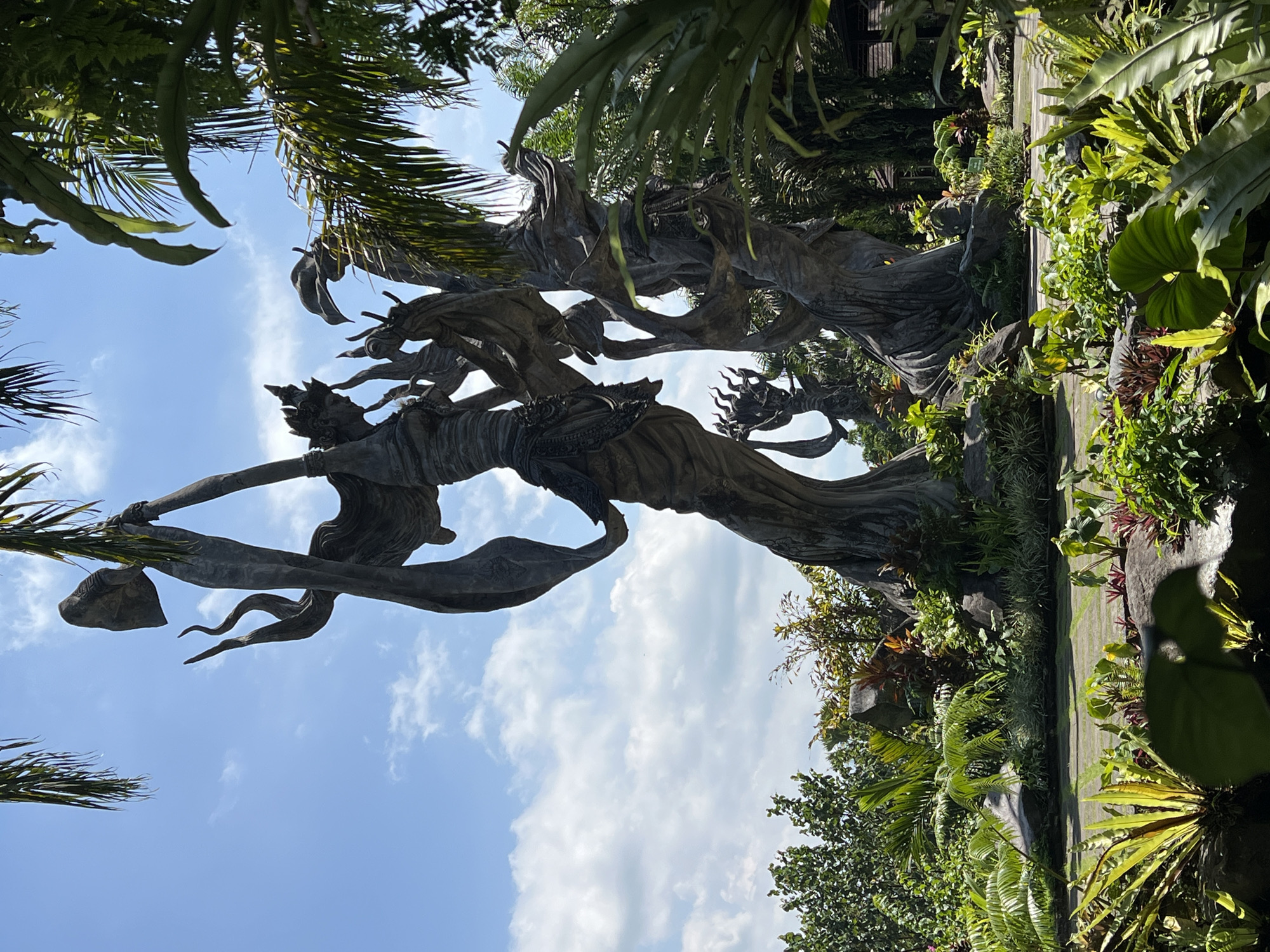 The image depicts a sculpted stone statue of a female figure, possibly a deity or mythological character, in a lush, tropical setting. The statue appears to be part of a landscaped garden, with a variety of plants and trees surrounding it. The sky is visible in the background, indicating an outdoor location. The statue is likely located in Kedewatan, Indonesia.