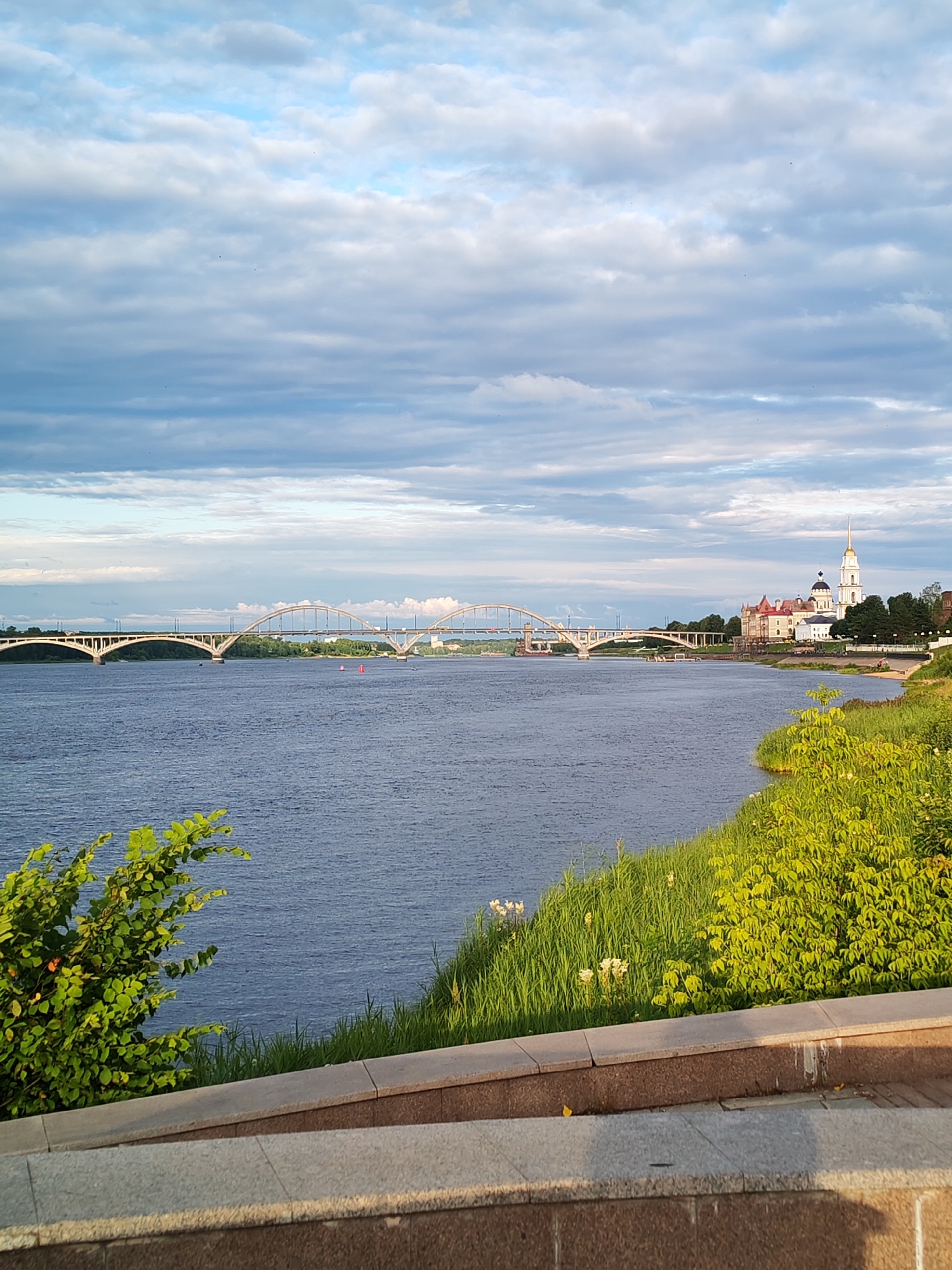 The image depicts a wide river in Bol'shie Mkhi, Russia, under a partly cloudy sky. A large, arched bridge spans the river in the middle ground, with several smaller structures and trees visible on the far bank. On the right bank of the river, a cluster of buildings, including a church with a tall spire, stands prominently. Lush green vegetation covers the riverbank in the foreground and midground, with a stone embankment in the immediate foreground. No people are visible in the image. The overall scene is serene and picturesque, characteristic of a riverside landscape.