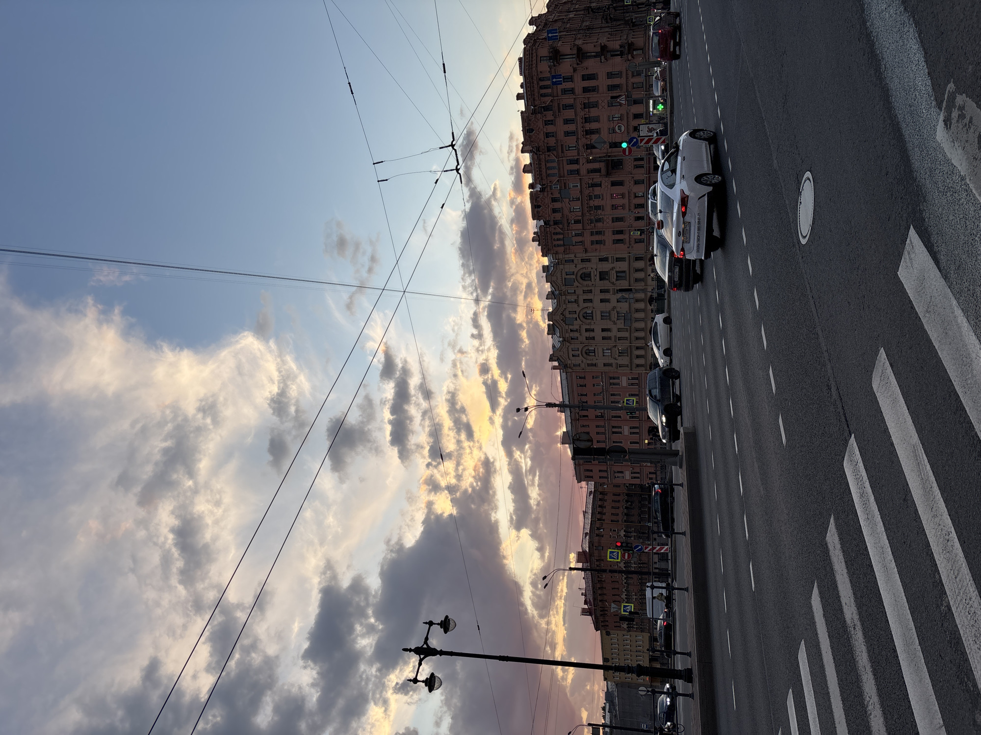 An outdoor, slightly rotated (appears as if portrait but taken in landscape), eye-level shot captures a bustling city street scene at what appears to be dusk or early evening in Saint Petersburg, Russia.
The upper portion of the image is dominated by a dramatic sky filled with scattered clouds, illuminated by warm, golden, and pinkish hues, suggesting a sunset. Numerous overhead wires, likely for trolleybuses or trams, crisscross the sky.
The middle ground features a long, multi-story building with reddish-brown or pinkish facades and numerous windows, stretching along the right side of the street. Its architecture appears classic European. Several utility poles with streetlights and traffic lights are visible along the sidewalk.
The foreground and lower right show a wide asphalt road with multiple lanes. White dashed lines mark the lanes, and a prominent pedestrian crosswalk with thick white stripes occupies the lower right corner. A circular manhole cover is visible in the middle of the road.
Several cars are present on the street: a white sedan is prominent in the middle right, with its rear visible, showing a partial license plate "X299..." Other cars, mostly dark-colored, are visible further down the road and parked.
Notable details include a green traffic light on the right, indicating active traffic flow in that direction, and a red traffic light further down the street on the left. Several road signs are visible: a blue sign with a left-pointing white arrow (likely one-way or turn instruction), a red "no entry" sign, and a blue pedestrian crossing sign with a white rectangular sign below it. The overall impression is an active urban environment transitioning from day to night.