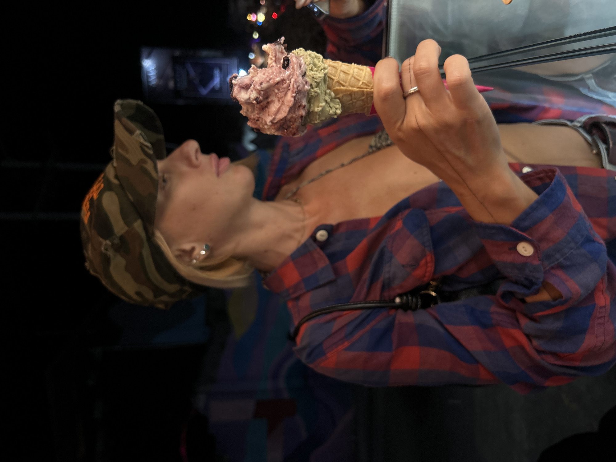 In Kuta Selatan, Indonesia, a woman wearing a camouflage baseball cap and a red and blue plaid shirt is holding a double-scoop ice cream cone. The ice cream has a green scoop at the bottom and a pink scoop with berries on top. She is also wearing a necklace and a ring. The background is dark and appears to be outdoors at night, with some blurred lights visible.