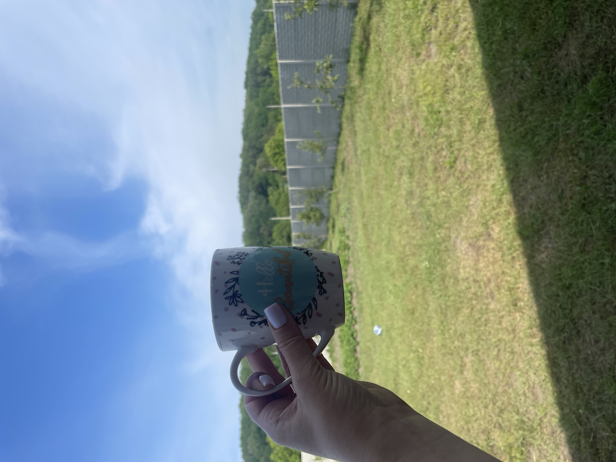 The image captures an outdoor scene in Petrivka, Ukraine, on a bright day. In the foreground, a left human hand, adorned with light lavender nail polish, holds a white ceramic mug. The mug features pink polka dots and a central teal circular design with green leaf motifs, containing the golden cursive text "Hello Beautiful." The ground beneath consists of a somewhat patchy green and dry brown grassy area, with a distinct dark shadow extending diagonally from the lower right corner. In the mid-ground, a lengthy, segmented grey concrete fence runs horizontally, behind which small green saplings are planted at intervals. The background reveals a continuous line of green forested hills under a clear blue sky dotted with soft white clouds, more concentrated towards the upper right.