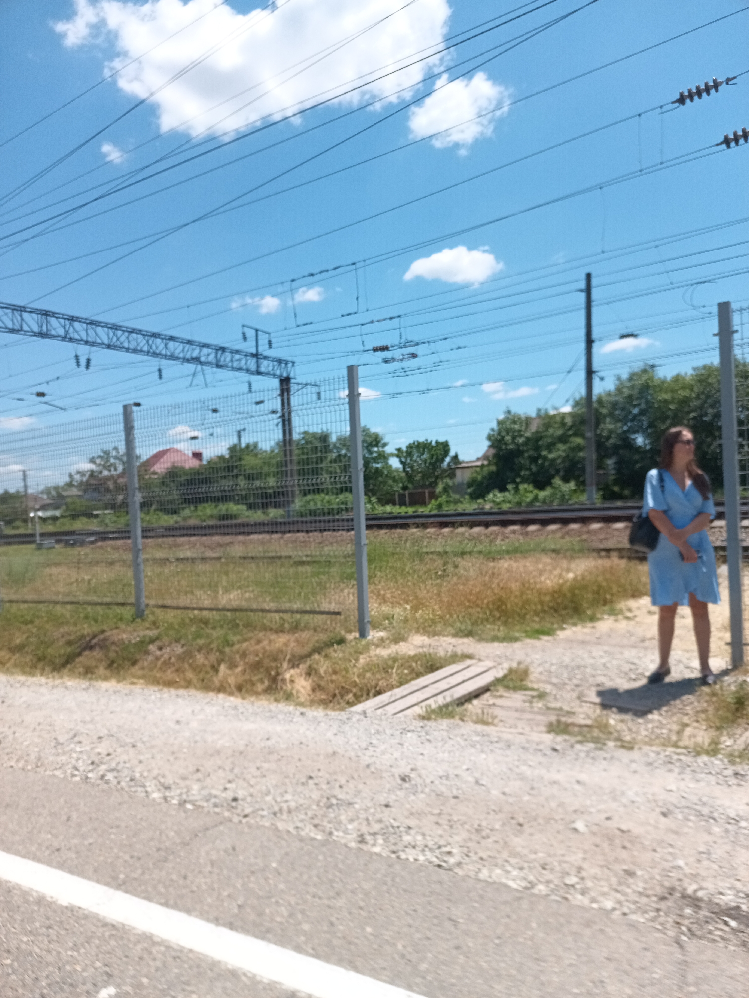 The image depicts a view of a railway track in Krymsk, Russia, under a clear blue sky with scattered white clouds. A young woman, wearing a light blue dress and carrying a dark bag over her shoulder, stands near a fence. She is looking to the right, away from the camera. The fence, made of metal mesh with grey posts, runs along the railway. Beyond the fence, railway tracks are visible, with sparse vegetation and trees in the background, including a house with a red roof. A road with a white line marking is in the foreground. The overall scene suggests a sunny day in a semi-urban or rural setting near a railway line.