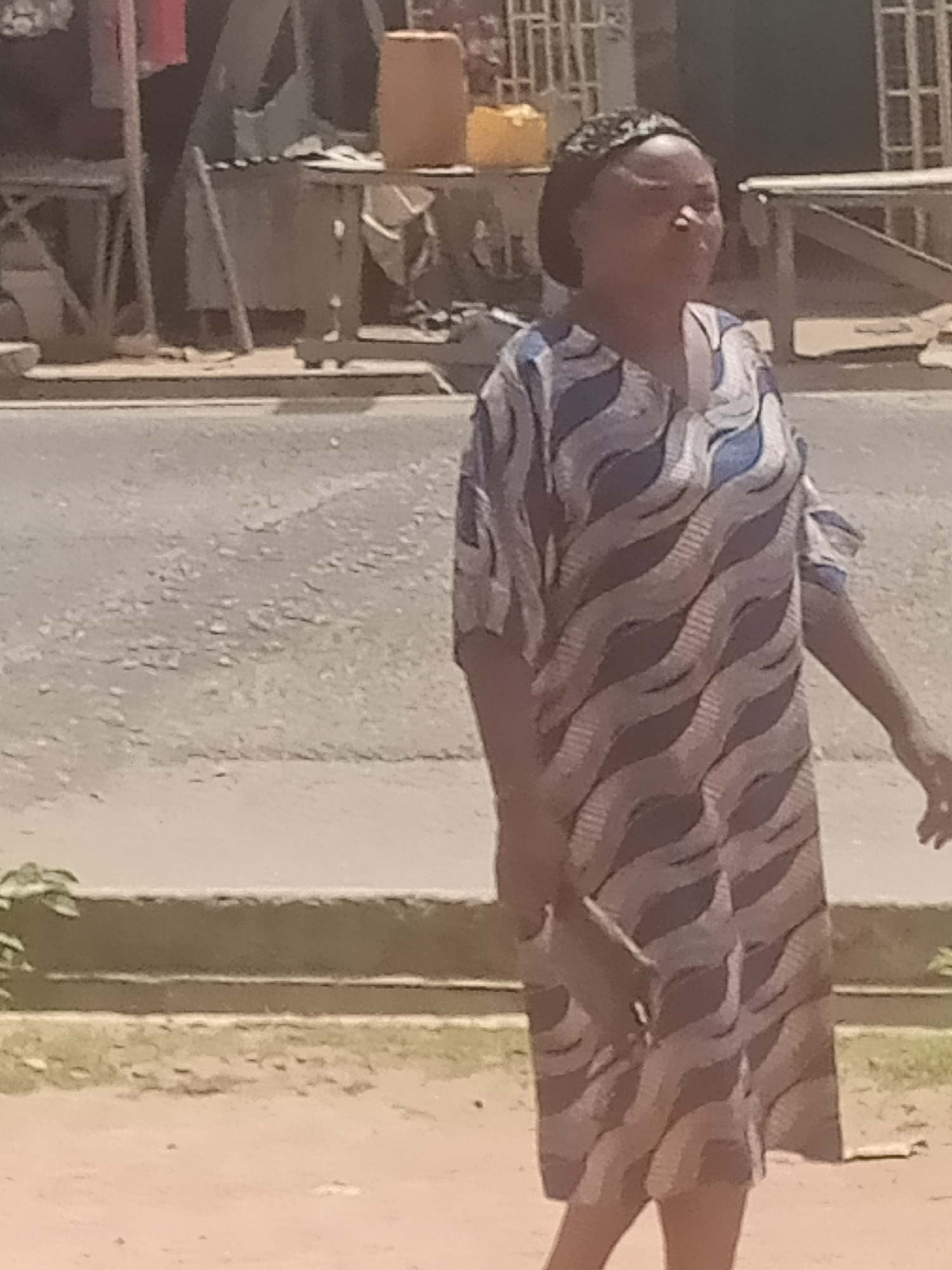 The image depicts a woman walking outdoors in Jalingo, Nigeria. She is wearing a patterned dress with wavy blue and gray stripes and appears to be holding a dark-colored object in her right hand. The background shows a street scene with what appears to be market stalls or shops. The ground is paved, with some vegetation visible. The lighting suggests it is daytime.