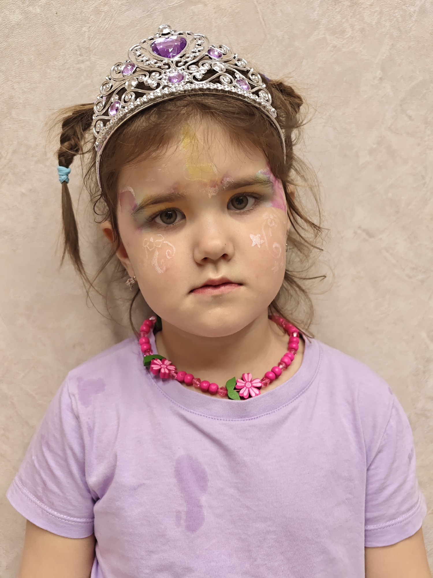 A young girl with face paint, a crown, and a necklace is standing indoors against a textured beige wall. She has brown hair styled in two small pigtails and is wearing a light purple t-shirt. The girl’s face is decorated with colorful makeup around her eyes and white designs on her cheeks. She has a silver crown adorned with purple jewels and a pink beaded necklace with flower accents. There is no indication of a specific location in Chaikovsky, Russia, in the image.