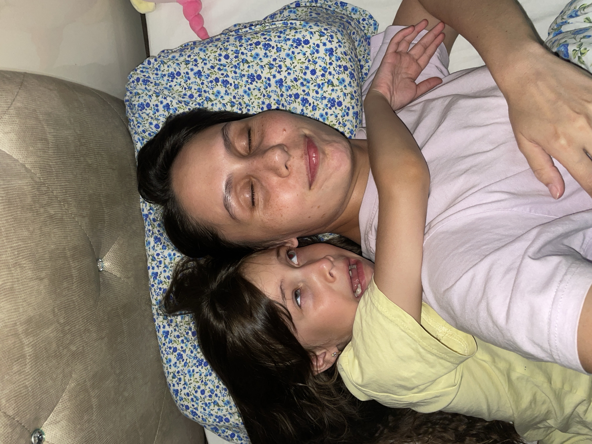 A young girl lies on a floral pillow next to an adult woman, both on a bed in an indoor setting. The woman is wearing a light pink shirt and has her eyes closed. The girl is wearing a yellow shirt and looking up at the camera. The headboard of the bed is upholstered in a beige fabric with diamond button detailing. There is a soft toy with pink and yellow coloring on the pillow above the woman. The location, Izhevsk, Russia, is irrelevant to the immediate, observable contents of the image.