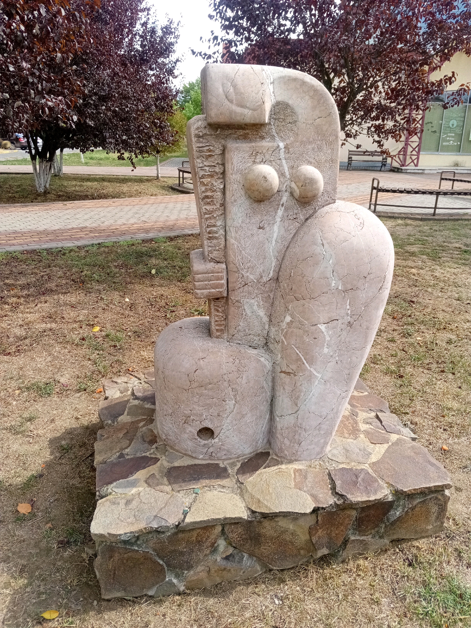 An abstract stone sculpture, likely representing a female form, stands on a stone-covered base in a park-like setting. The sculpture is composed of light-colored, veined stone and features rounded shapes suggesting breasts and a seated posture. Behind the sculpture, a paved walkway with a reddish-brown brick pattern is visible, bordered by dry grass. Several trees with dark reddish-brown foliage are present in the background, along with benches and a building with windows. The scene is outdoors, under natural lighting, suggesting it is daytime. The location is identified as Tiachiv, Ukraine.