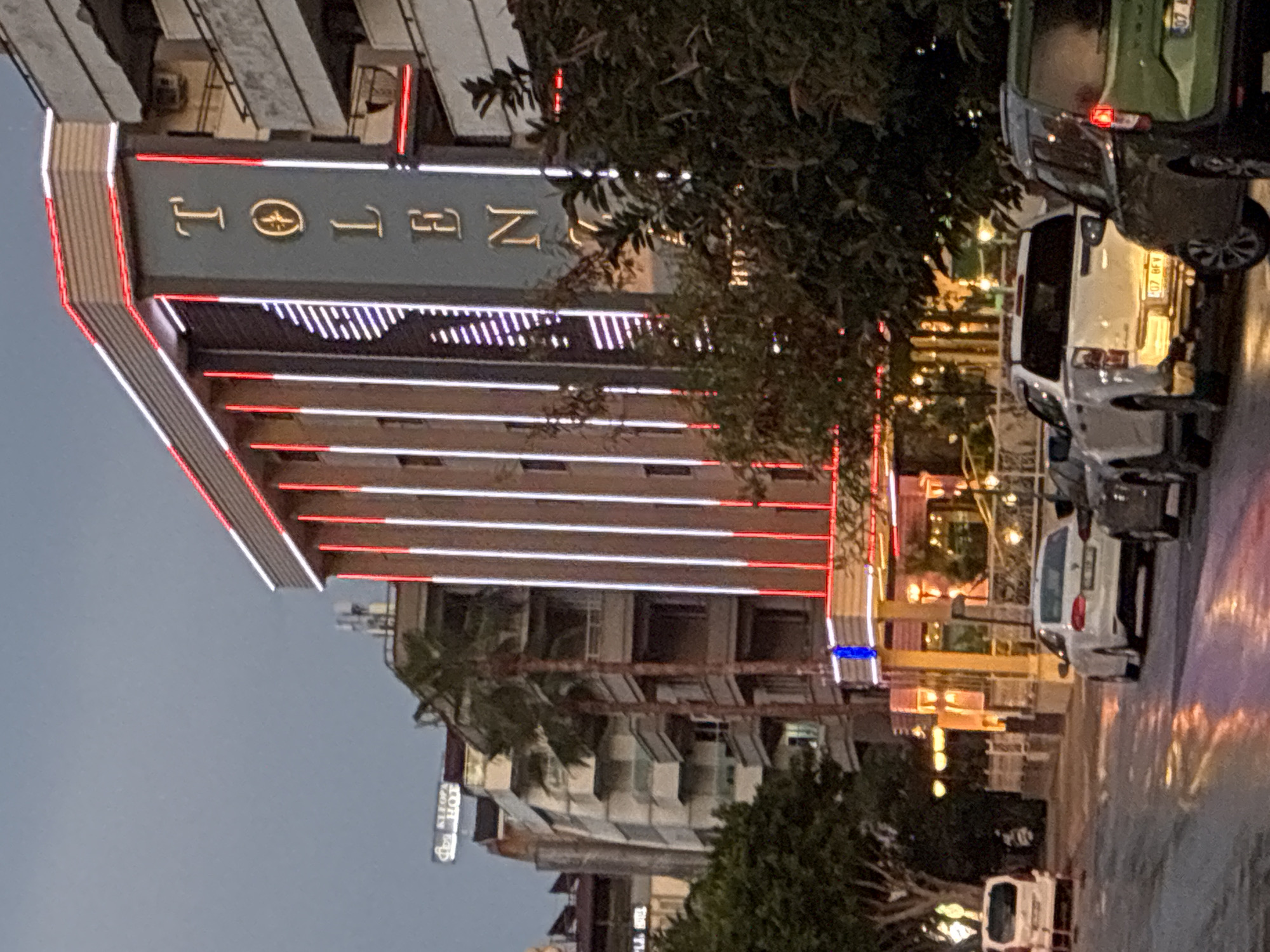 This image captures an urban street scene in Alanya, Turkey, at twilight or evening, characterized by artificial illumination.
The focal point is a prominent multi-story building on the right, featuring a dark grey-blue facade. Vertically displayed in gold lettering on this facade is the word "TOLEN," with the 'O' stylized as a circular, compass-like emblem. The building is extensively lit with vertical red and white LED strips running up its height, and a continuous red LED strip outlines the perimeter of its roof. A bright blue light accent is visible near the base of the building's facade.
To the immediate right of the "TOLEN" building, an older, multi-story building with numerous balconies is partially visible. Further down the street, to the left of the "TOLEN" building, another multi-story structure features a rooftop sign partially reading "KLEOPA HOT," suggesting a "Kleopatra Hotel." Palm trees and other leafy vegetation are interspersed between and in front of the buildings, lining the street.
The asphalt road in the foreground is visibly wet, reflecting the colorful artificial lights from buildings and vehicles in shimmering patterns of orange, red, and white. Several vehicles are parked or moving on the road:
* In the foreground on the right, the rear of a dark green SUV or minivan is visible, with a partial license plate starting "07 AFV."
* Behind it, the rear of a white pickup truck is clearly visible, bearing the license plate "07 BFV 90."
* Further down the street on the left, a white sedan's rear is visible.
* On the far left, a white van is parked.
The license plates beginning with "07" confirm the location within Antalya Province, where Alanya is situated. No individuals are discernible within the frame.