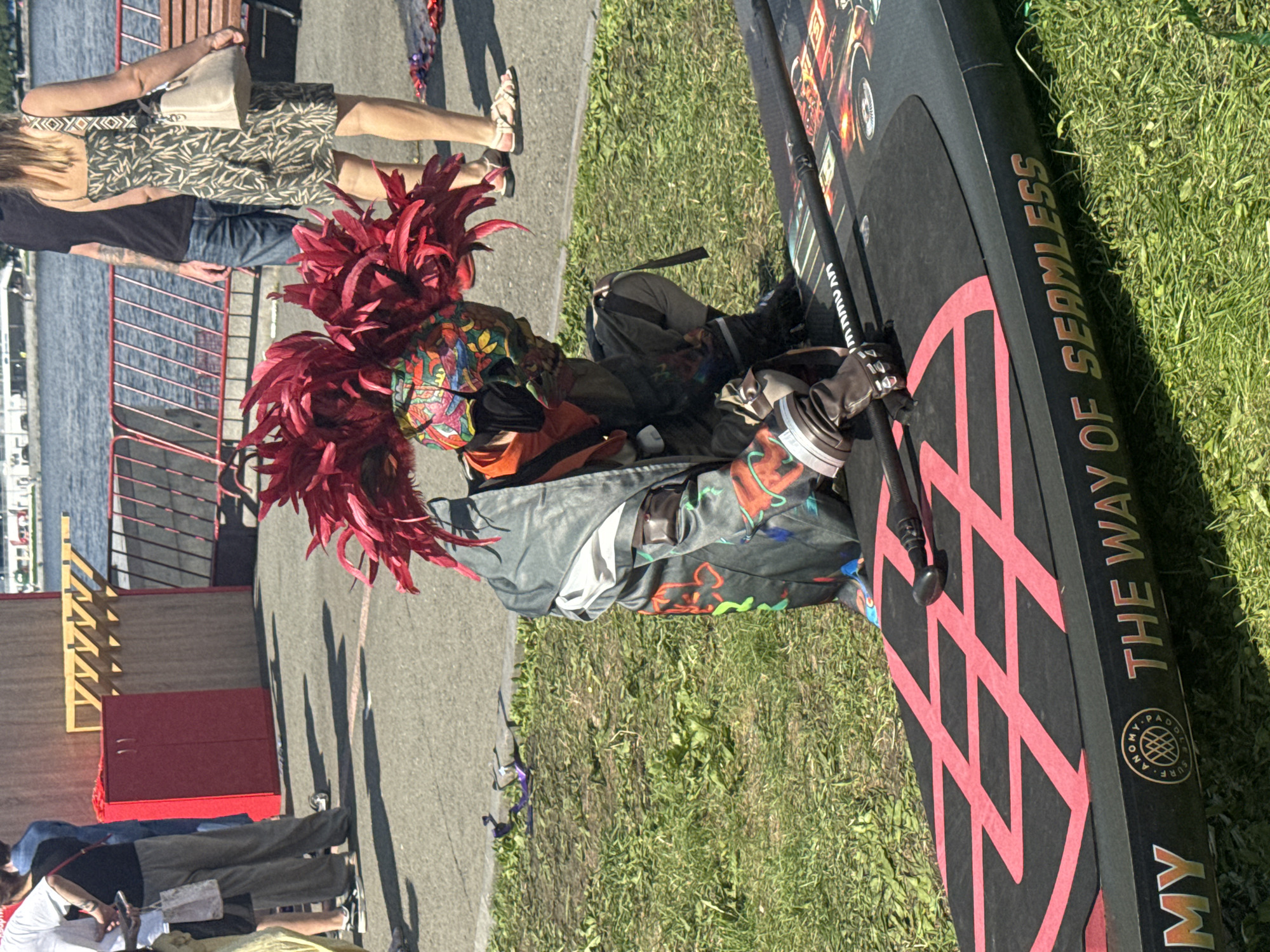 A person in a red feathered headdress and a colorful costume is kneeling on a paddleboard. The paddleboard has "THE WAY OF SEAMLESS" printed on its side. The scene appears to be outdoors, possibly near a body of water, with grass and pavement visible. The background shows a railing, a red cabinet, and people in the distance. The lighting suggests it is daytime. The location cannot be determined from the image alone.