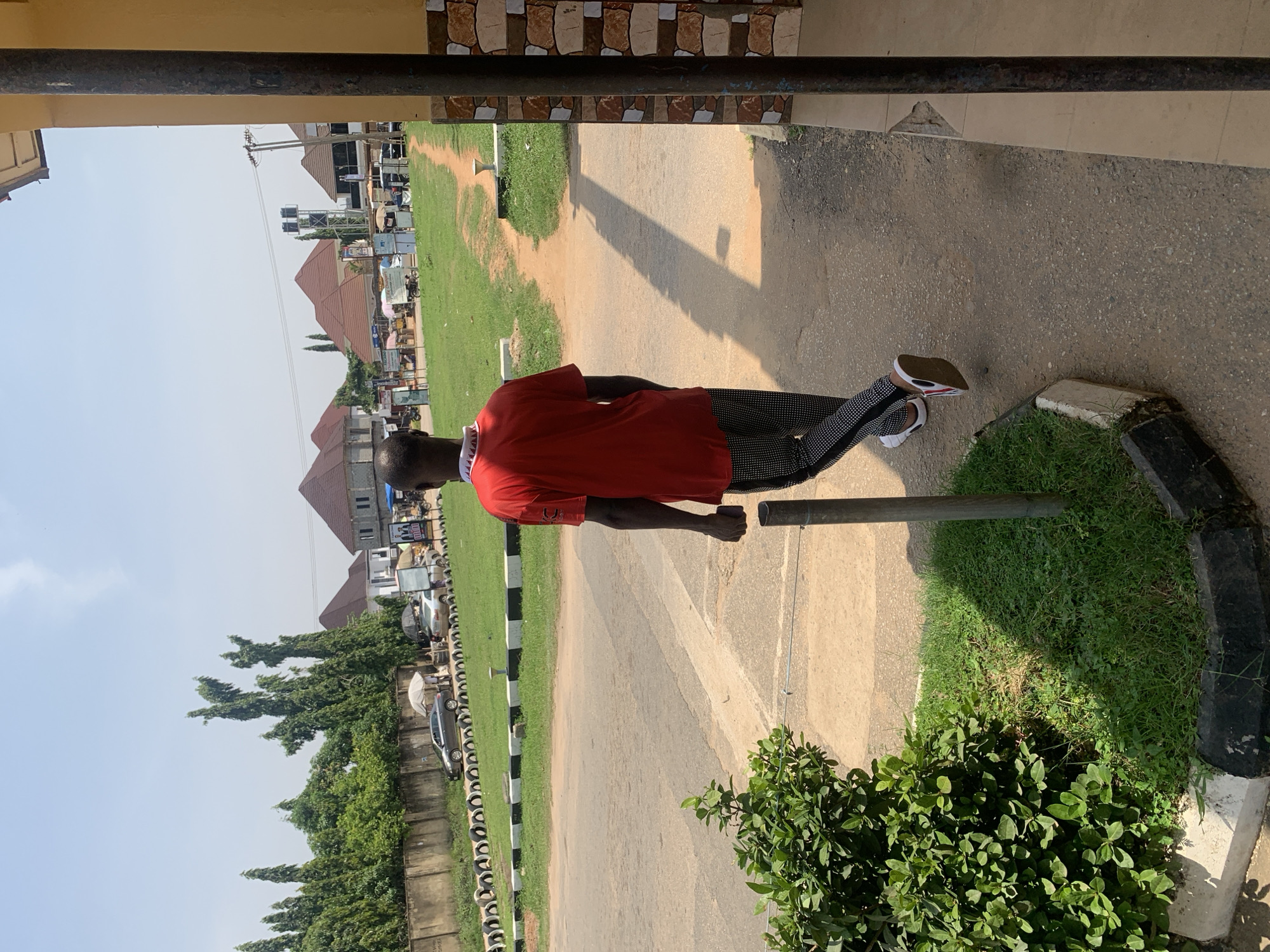 The image depicts an outdoor scene on a sunny day, likely in Abuja, Nigeria, given the architectural styles and vegetation. A young Black male, viewed from behind, is walking on a paved pathway next to a road. He is wearing a red t-shirt and black pants with a white patterned design. His feet are clad in white sneakers with red and black accents. To his left, there is a vibrant green bush and a patch of grass bordered by a curb painted black and white. A metal pole stands in the grassy area. Beyond the pathway and a green lawn, a dirt path leads towards a street lined with buildings, some of which have distinctive red, conical roofs. Several cars are parked along the side of the road, and various stalls or small shops are visible, suggesting a commercial or market area. Tall trees with sparse foliage are scattered throughout the background. The sky is clear blue with a few wispy clouds. The overall impression is of a somewhat bustling urban environment.