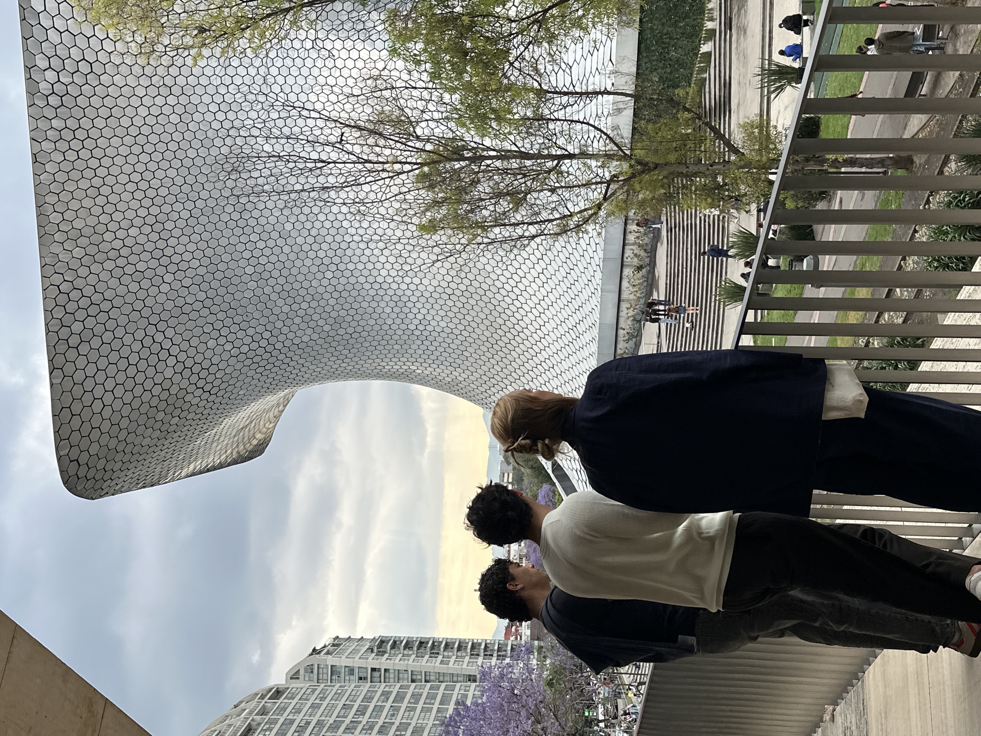 The image is taken outdoors, likely on a pedestrian bridge or elevated walkway in Mexico City, Mexico. Three people are standing side-by-side, their backs to the camera, looking towards a unique, curved building with a distinctive hexagonal patterned facade. The architecture suggests that this is the Museo Soumaya in the Nuevo Polanco district.
The person closest to the camera is wearing a dark blue jacket, while the second person is wearing a white shirt, and the third person is wearing a dark grey shirt. The walkway is bordered by a metal railing and a glimpse of green space, possibly a park, is visible beyond it. In the background, a modern building with numerous windows and a tree with purple flowers are visible. The sky is overcast. Other people are visible walking in the background, on steps and pedestrian paths.