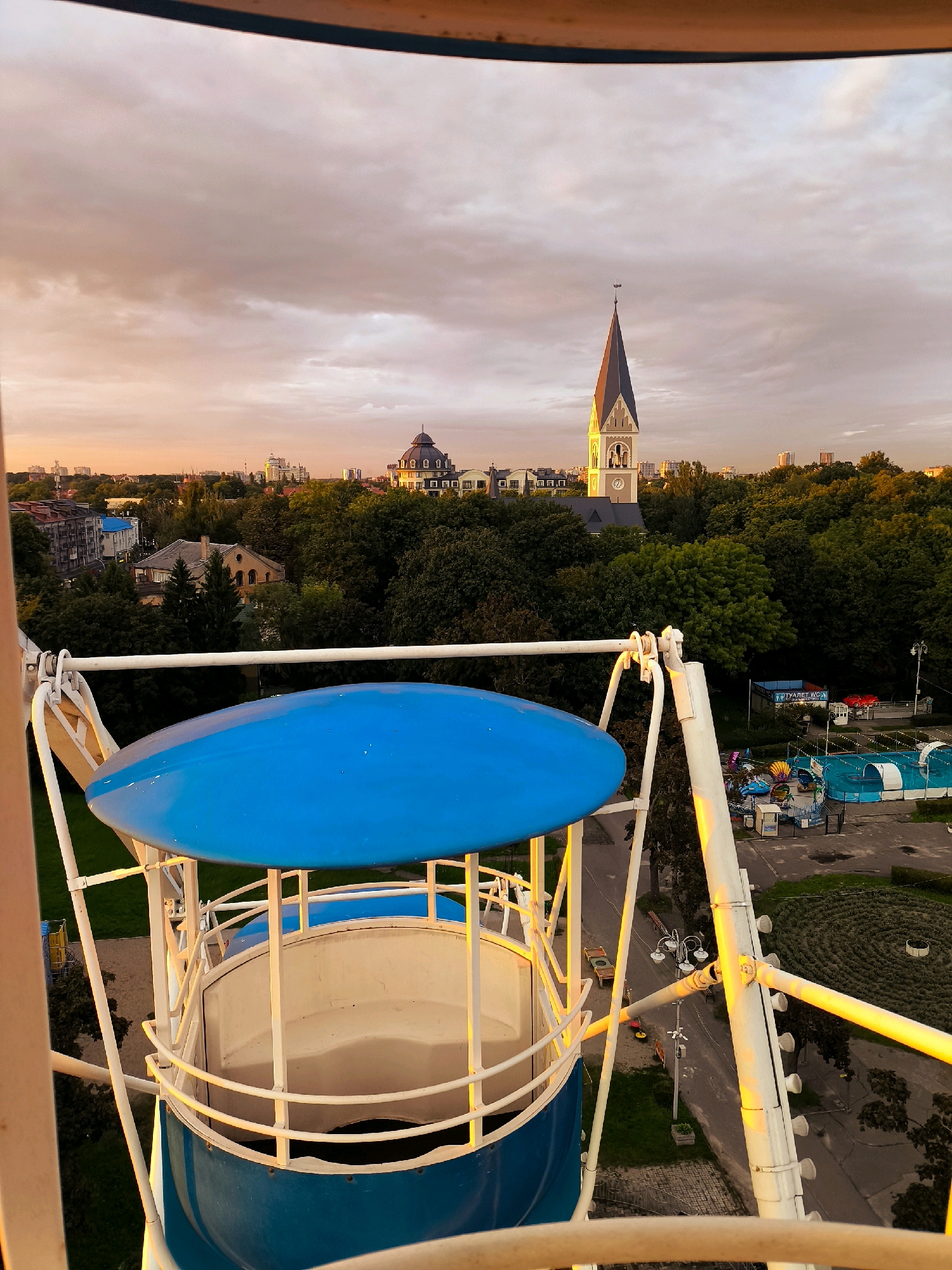An elevated view from a Ferris wheel gondola shows a park and cityscape during a sunset. The gondola, painted blue and white, dominates the foreground. In the mid-ground, a tall, pointed church steeple with a clock is visible amidst dense green trees. Beyond the trees, buildings and the orange-tinted sky are seen. To the right, an amusement park with blue water attractions and smaller rides is visible. The scene suggests the amusement park is located in a city with parks and historical architecture, characteristic of Kaliningrad, Russia.