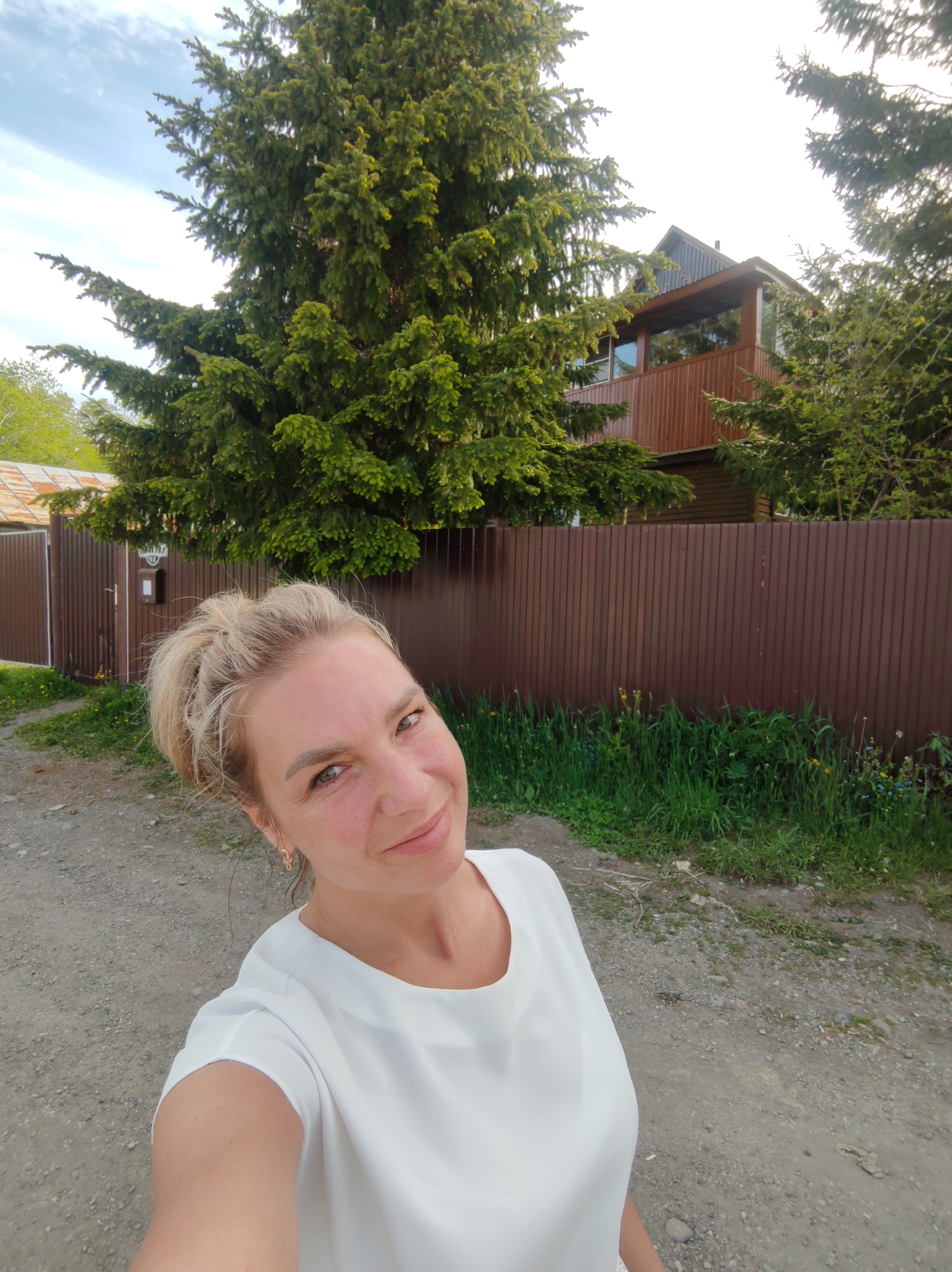 A woman with blonde hair pulled up into a messy bun, wearing a white shirt, takes a selfie in front of a brown corrugated metal fence. Behind the fence, a two-story wooden house is visible through the branches of a large green spruce tree. The ground is covered in gravel, with patches of green grass and small wildflowers growing along the fence. A metal gate is visible to the left of the woman. The sky above is blue with scattered white clouds. This scene is located in Paratunka, Russia.