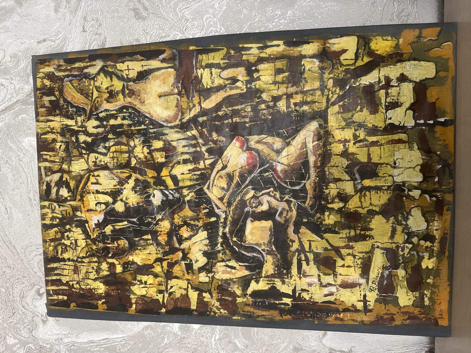 The image shows an abstract painting on a canvas, possibly acrylic or oil, mounted on a wall. The color scheme is primarily gold, brown, and black, with a textured appearance resembling brickwork or stone. The painting includes discernible figures, appearing to be human forms in a partially obscured arrangement, suggesting a narrative or symbolic scene. In the lower right corner, there's a signature "B.Pilauri" written. The painting is surrounded by a dark grey frame. The wall behind the painting has a light-colored, patterned wallpaper, which adds a sense of domestic or interior setting. There is no direct evidence to determine its location to be in Tbilisi, Georgia.