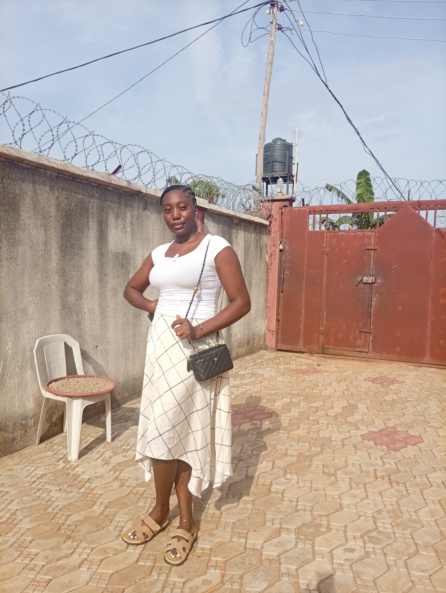 A full shot captures a young Black woman standing in a paved courtyard in Unknown, Unknown. She is dressed in a short-sleeved white top and a white plaid skirt. A black purse hangs from her right shoulder, and she wears light brown sandals. She is posing with her right hand on her hip.
Behind her is a gray wall topped with barbed wire, a rusted maroon metal gate, and a black water tank on a tall concrete pillar. An electrical pole with wires extending out of the frame is visible in the background. To her left is a white plastic chair with a flat, woven circular tray on the seat. The paving stones underfoot are light brown with red accents. The sky is light blue with faint clouds.