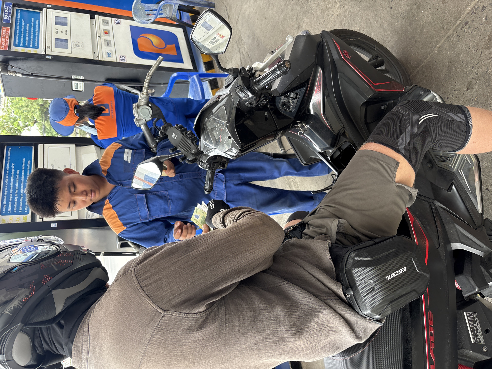 A man on a motorcycle is paying a gas station attendant at a Petrolinex gas station. The gas station attendant, dressed in a blue and orange uniform, is holding Vietnamese currency in his hand. The man on the motorcycle is wearing a helmet and grey pants. A black motorcycle with a black TAKEZERO bag and knee pad is positioned between the man and the gas station attendant. The gas station has a blue fuel pump, and a sign with information is displayed. OCR data from the image identifies Xăng E10 RON95 and DO 0.05S-II fuels. The location is identified as Bac Nha Trang Ward, Vietnam.
