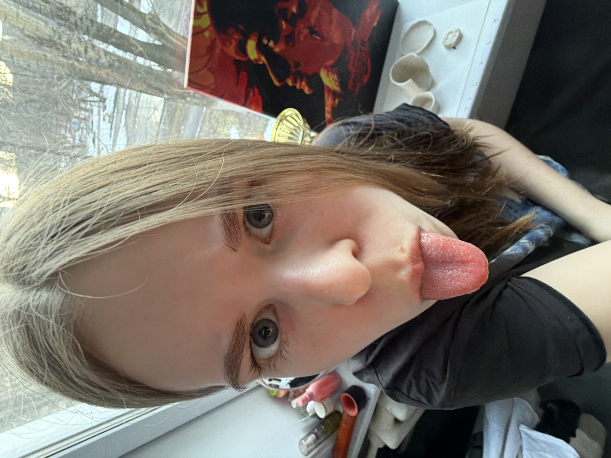 A blonde-haired person, presumably female, extends their tongue towards the camera. The background shows a window with trees outside, a picture of a man with red/orange tones, and what appear to be white ceramic objects. A container with various bottles and tubes sits adjacent to the person, implying an indoor setting. The exact location is not evident from the image.