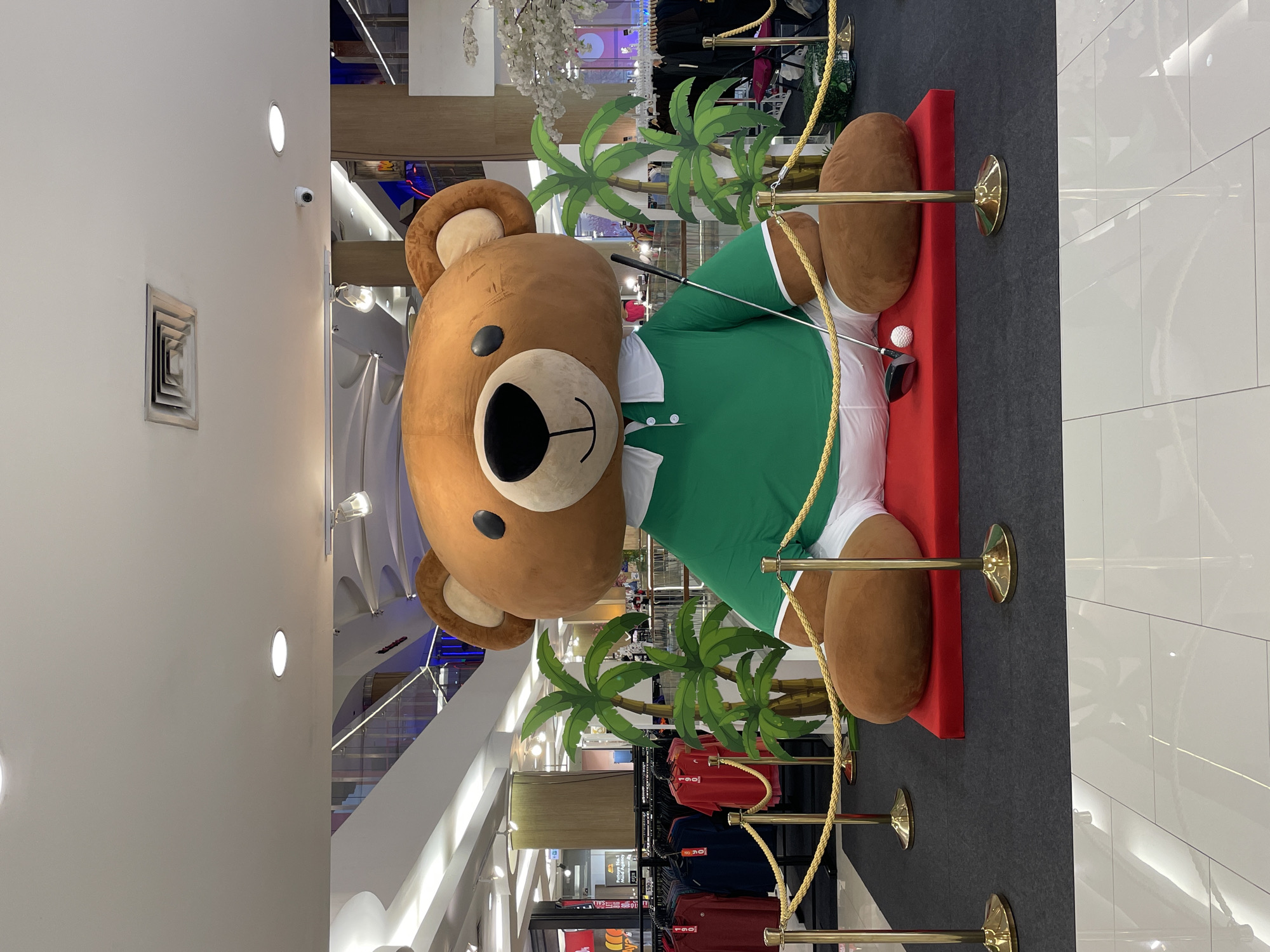 A large, inflatable teddy bear is displayed in a shopping mall. The bear is wearing a green polo shirt with a white collar and cuffs, and white shorts. It is sitting on a red platform, with a golf club and ball placed in front of it. Two artificial palm trees are positioned behind the bear. The background shows racks of clothing for sale and escalators. The floor is tiled with a reflective white surface. The overall scene suggests a promotional display or event within a retail environment in Pattaya City, Thailand.
