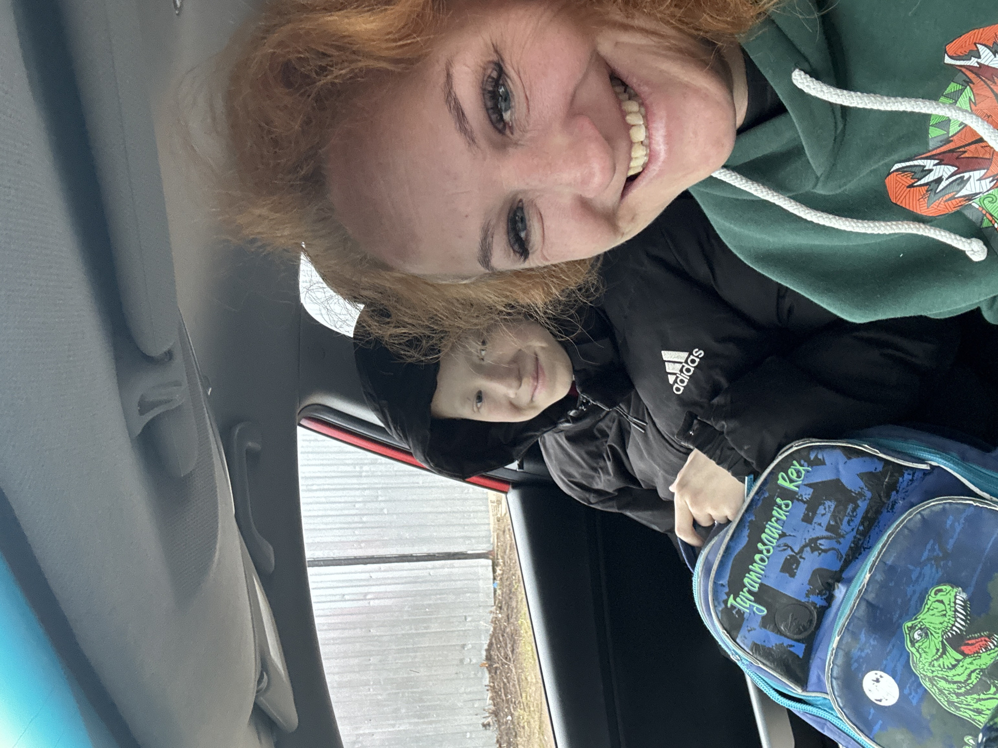 Two people are in a vehicle. The driver is a woman with red hair, wearing a green hooded sweatshirt. The passenger is a child wearing a black jacket with the Adidas logo. The child has a blue dinosaur-themed backpack. The view outside the window shows a fence. The location is not specified or inferable from the image. The specific city of Chaikovsky, Russia, cannot be verified based on the image.