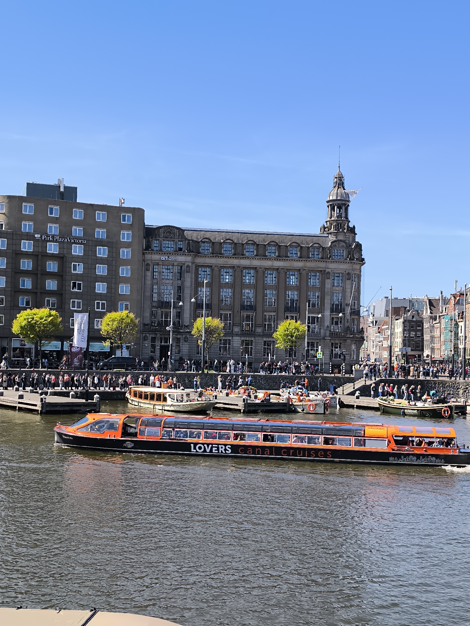 The image depicts a canal scene in Amsterdam, Netherlands, on a clear day. A "Lovers Canal Cruises" tour boat, predominantly black and orange, is moving across the water, filled with passengers visible through its windows. Along the canal banks, crowds of people are walking, standing, and interacting. Various smaller boats are docked or moored.
Buildings form the backdrop. On the left, there is a multi-story building, possibly a hotel called "Park Plaza Victoria", followed by a grander, older building with a prominent tower. Further buildings in various architectural styles line the canal. The sky is blue and clear. The water is choppy, reflecting light and images of the surroundings.