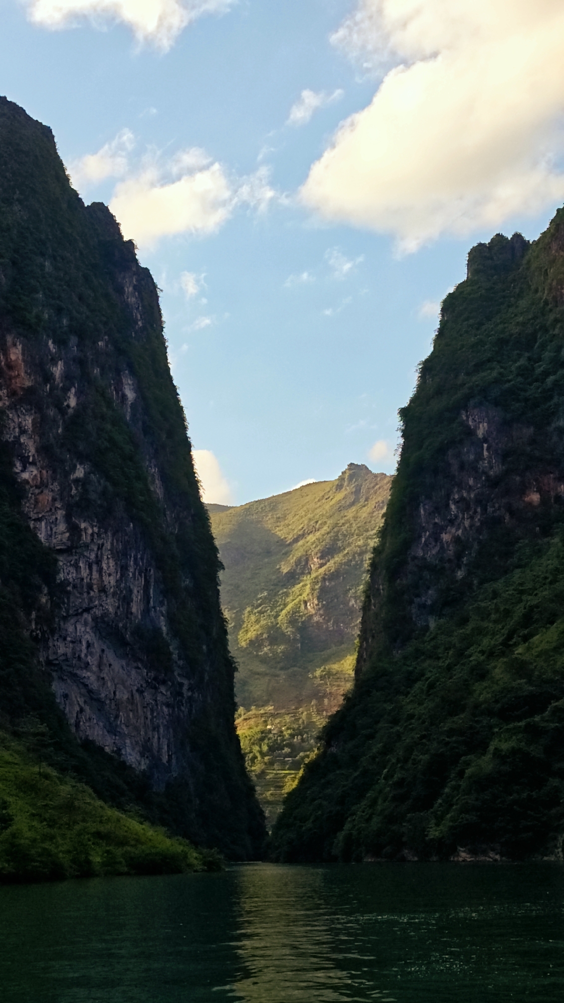The image shows a narrow river valley in Đồng Văn Commune, Vietnam. Towering, verdant mountains flank both sides of the river, with a sliver of blue sky and scattered clouds visible above. The mountains exhibit varied textures, from sheer rock faces to dense vegetation. The water reflects the light and shadows, appearing a deep green color. The light in the distance illuminates terraced fields or small settlements.