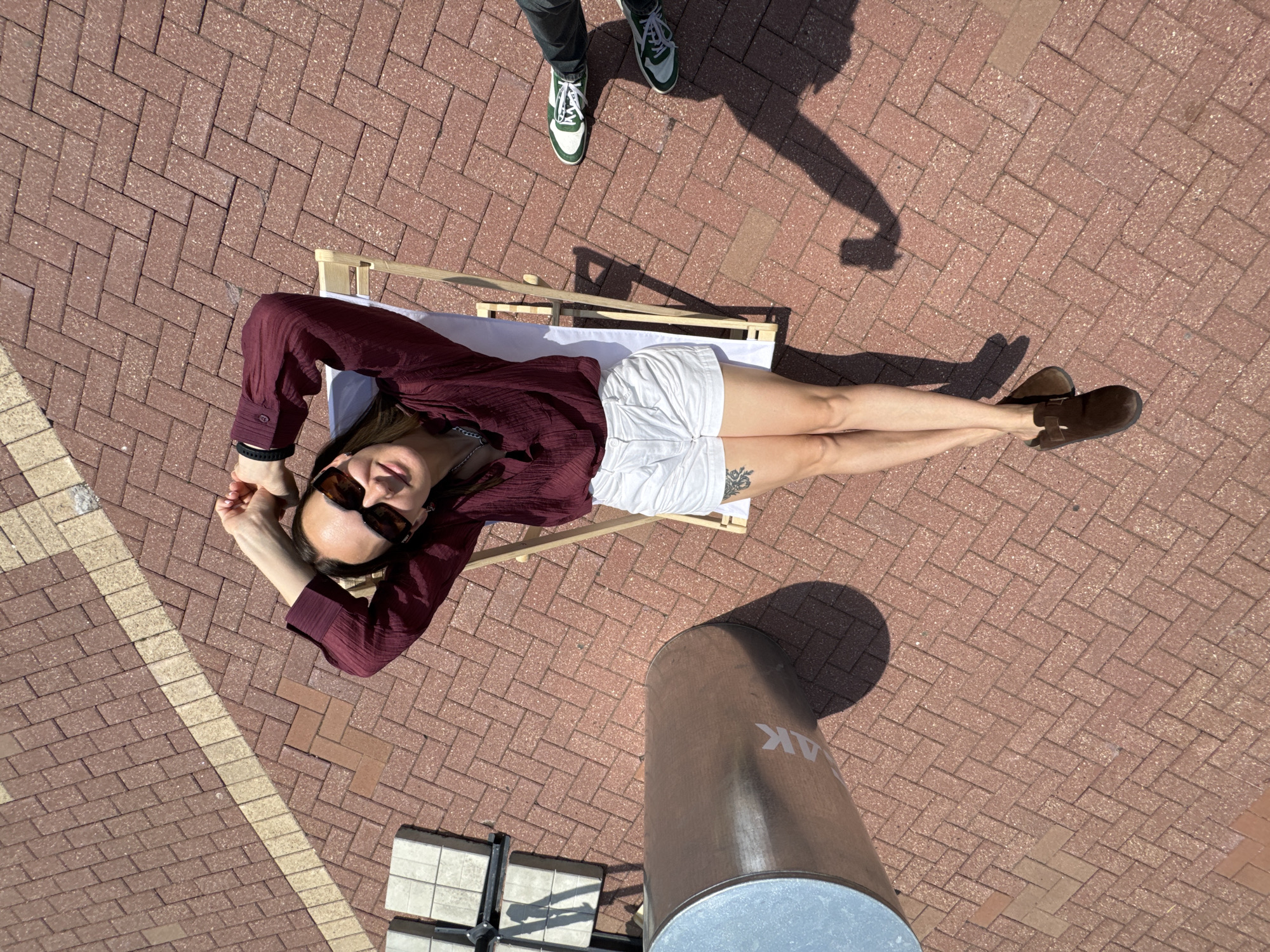 In Estosadok, Russia, on a sunlit day, a young woman reclines on a wooden beach chair outdoors. She is wearing a dark maroon long-sleeved top, white high-waisted shorts, and brown slip-on shoes. Her arms are raised behind her head, and she wears sunglasses. She has a tattoo on her left thigh. The ground is paved with reddish-brown bricks laid in a herringbone pattern. To her left is a large cylindrical silver object with the letters "AK" visible on its side. To her right, another person's legs are visible, wearing dark pants and green and white sneakers. Shadows from the woman, the chair, and the cylindrical object are cast on the brick ground. A white object, possibly a table, is partially visible in the upper left corner.