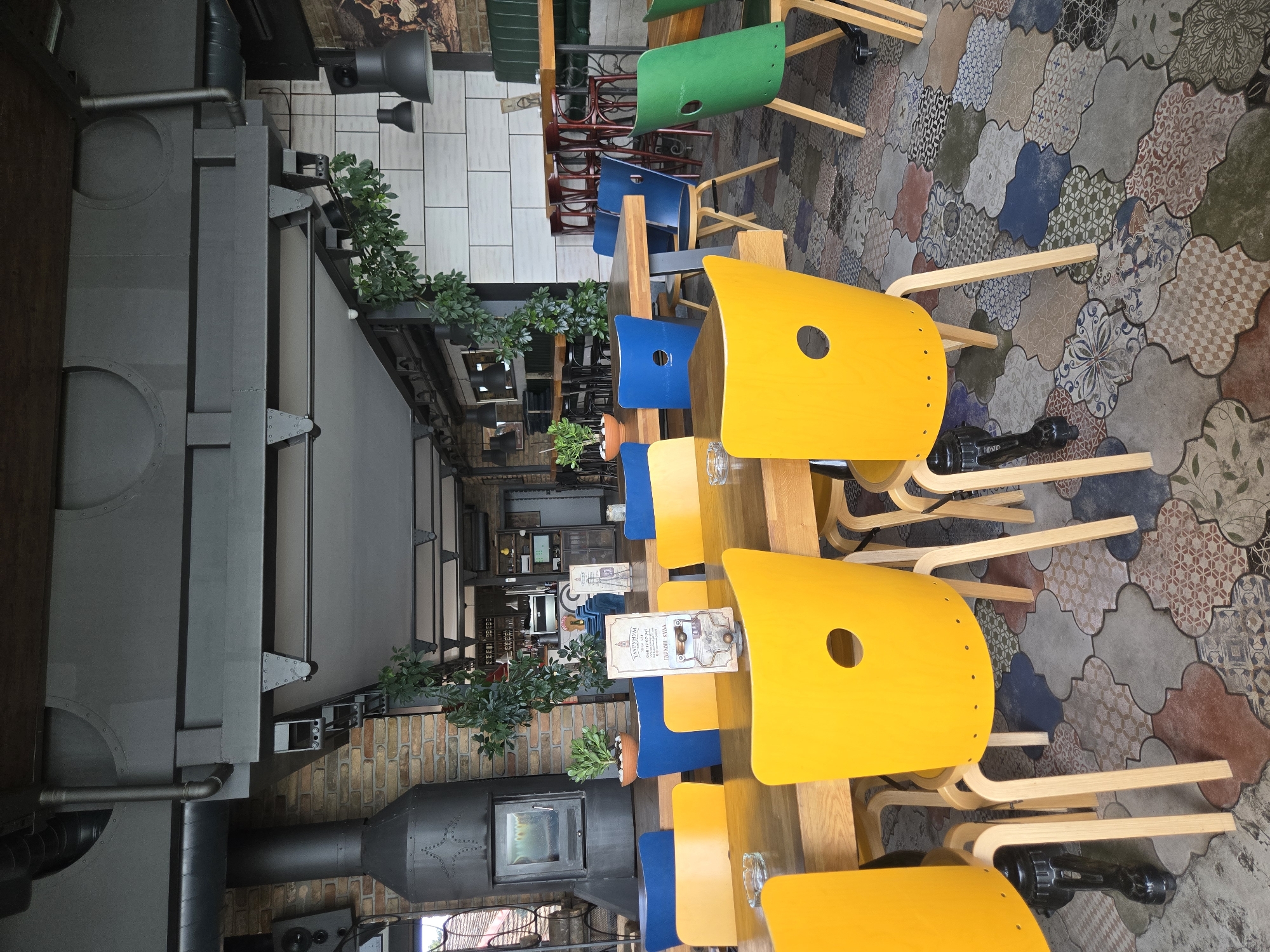 The image shows the interior of a cafe or restaurant in Zemun Urban Municipality, Serbia. A long wooden table is lined with a series of colorful chairs, alternating between yellow and blue, with a green chair visible further down the table. The floor is tiled with a pattern of irregular, variously colored and decorated shapes. Adjacent to the table, on the left side, is a large, black, possibly industrial-style structure, which may be part of the building's architecture or part of the restaurant's design. Green plants are placed on shelves and along the walls, adding a touch of greenery to the interior. The overall atmosphere suggests a modern, eclectic style within a seemingly industrial space.