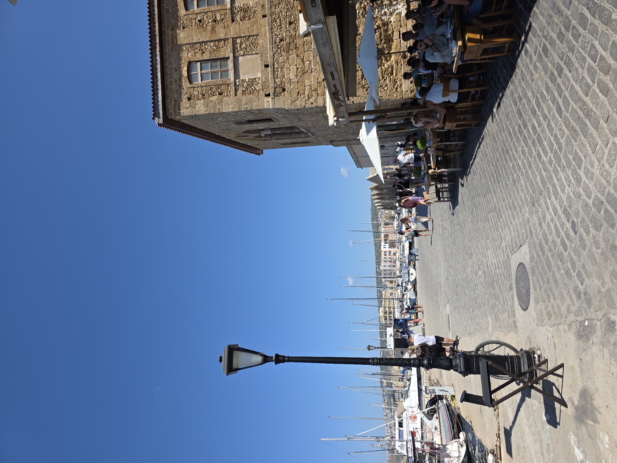In Chania, Greece, a sun-drenched cobblestone promenade runs alongside a harbor filled with sailboats. To the right, a stone building with arched windows stands, featuring an outdoor cafe with patrons seated at tables under umbrellas. A lamppost stands on the left side of the promenade, near a folded chair and a bicycle. People are walking along the promenade and dining at the cafe. The sky is a clear, bright blue.