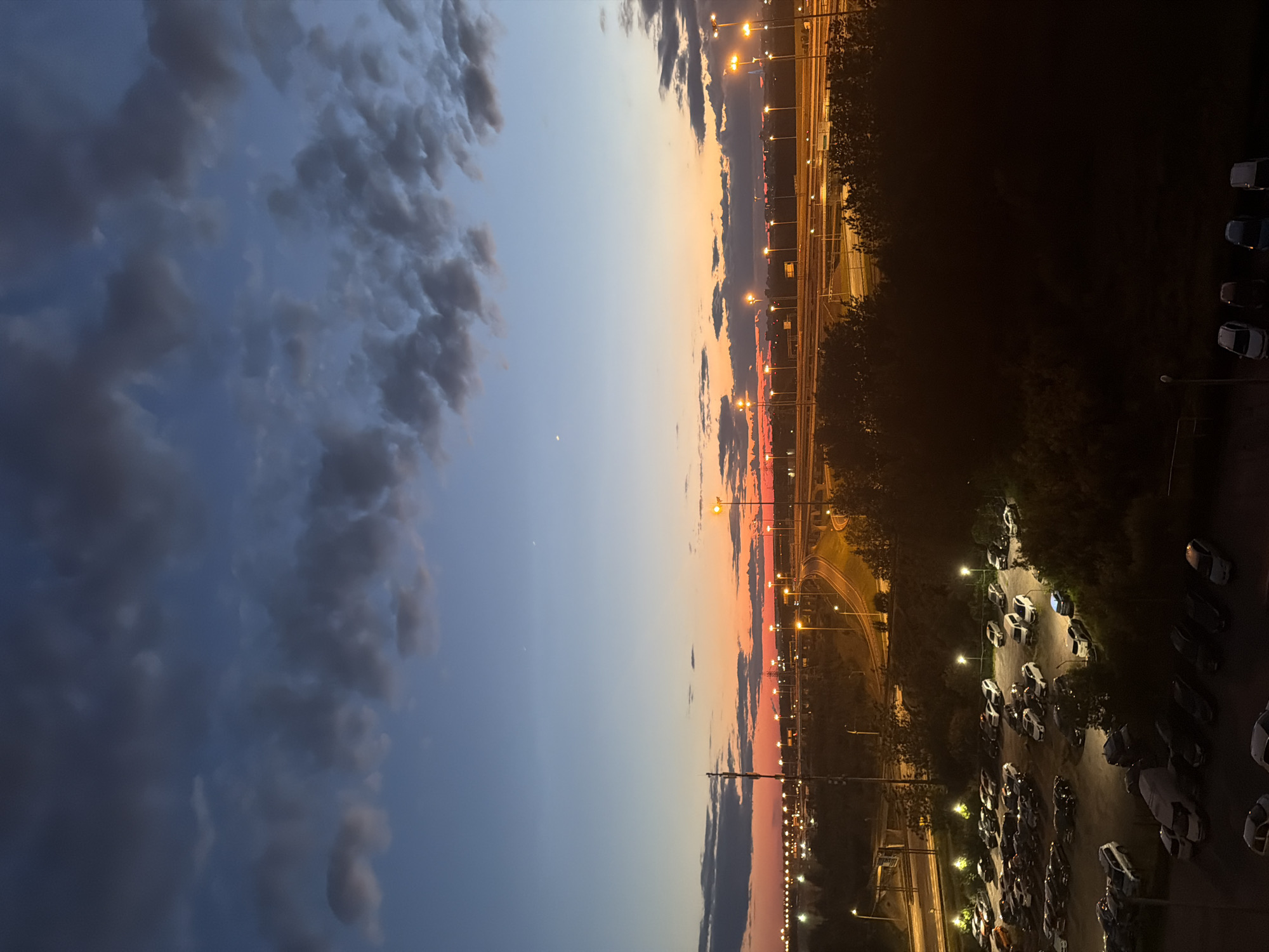 A high-angle, wide shot captures a city scene at dusk. The sky transitions from a deep blue at the top to warm orange and pink hues near the horizon, with scattered dark clouds. In the foreground, a dimly lit parking lot is filled with numerous cars, some illuminated by overhead lights. Beyond the parking lot, trees create a dark silhouette. In the midground, a highway with multiple lanes curves through the landscape, lined with streetlights casting pools of yellow light. In the background, a sprawling city skyline is visible, with more lights dotting the distance, suggesting a metropolitan area. The overall atmosphere is calm and transitional, marking the shift from day to night.