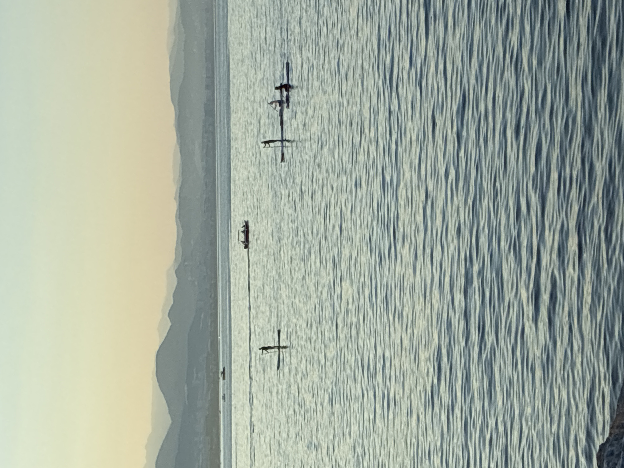 The image captures a serene scene of Alanya, Turkey, with a vast expanse of water dominating the foreground. The water, with its gentle ripples reflecting the soft light of what appears to be either dawn or dusk, is a tranquil blue-green hue. In the middle ground, several individuals are engaged in stand-up paddleboarding. One lone paddleboarder stands silhouetted against the water, paddle in hand. Further to the right, two more paddleboarders are visible, one paddling and the other seated on their board. In the distance, a small boat cruises across the water, leaving a faint wake. The background is characterized by a chain of layered mountains, their silhouettes softened by the atmospheric haze, fading into the pale, gradient sky. The sky transitions from a pale yellow near the horizon to a soft, light blue overhead. The overall impression is one of peacefulness and outdoor recreation at the scenic coast of Alanya.