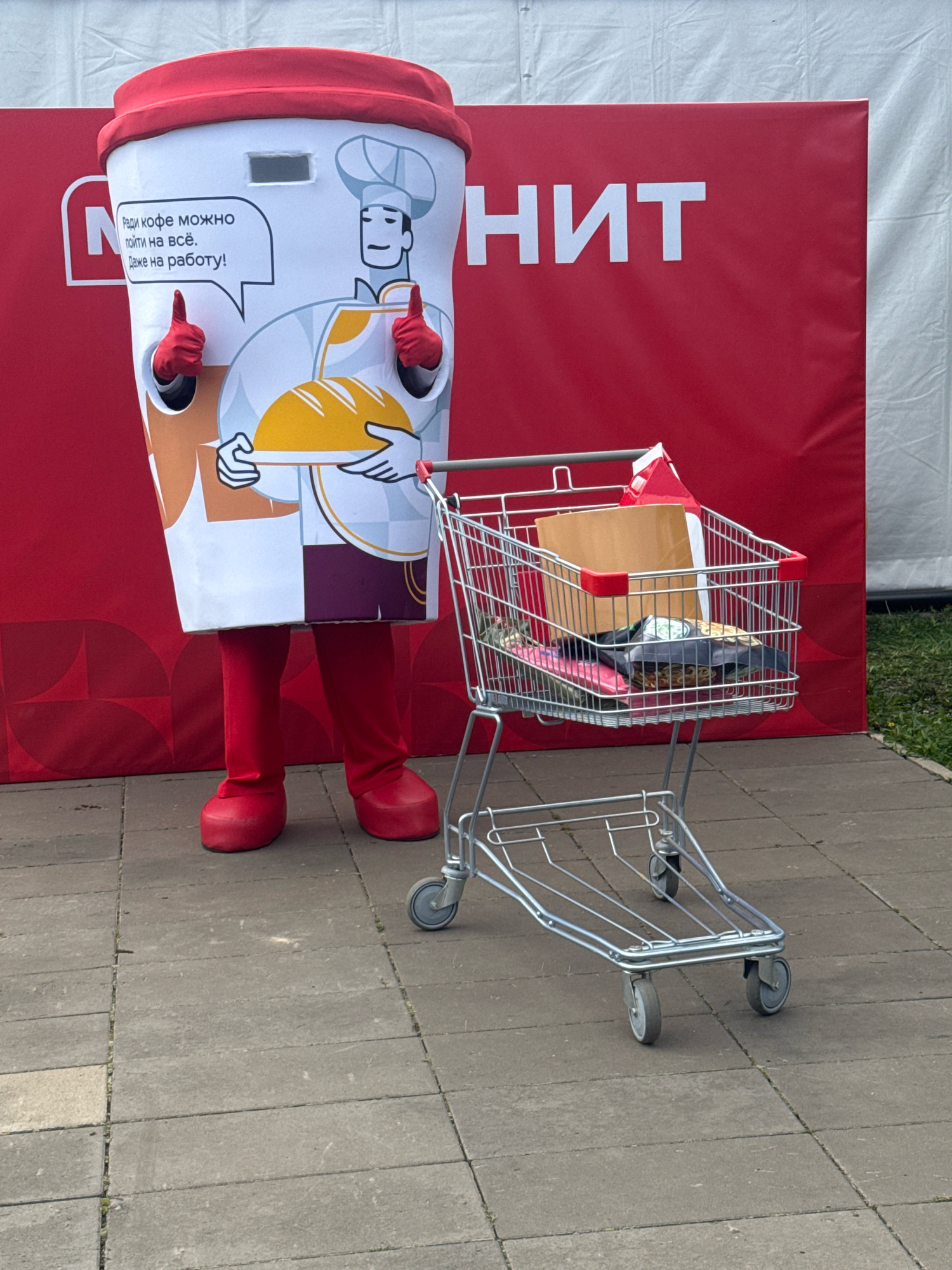 A mascot costume resembling a large coffee cup with arms and legs, wearing a chef's hat and apron and holding a loaf of bread, stands on a tiled sidewalk in front of a red backdrop with white text. The costume has a speech bubble that reads "Ради кофе можно пойти на всё. Даже на работу!" (For coffee, you can do anything. Even go to work!). Next to the mascot is a shopping cart filled with various items, including a cardboard box, bags, and other products. The background appears to be an outdoor event or promotional setup, possibly in Ufa, Russia. The mascot is wearing red gloves and boots, and its visible arms are giving a thumbs-up. The overall scene suggests a promotional event related to coffee or food.